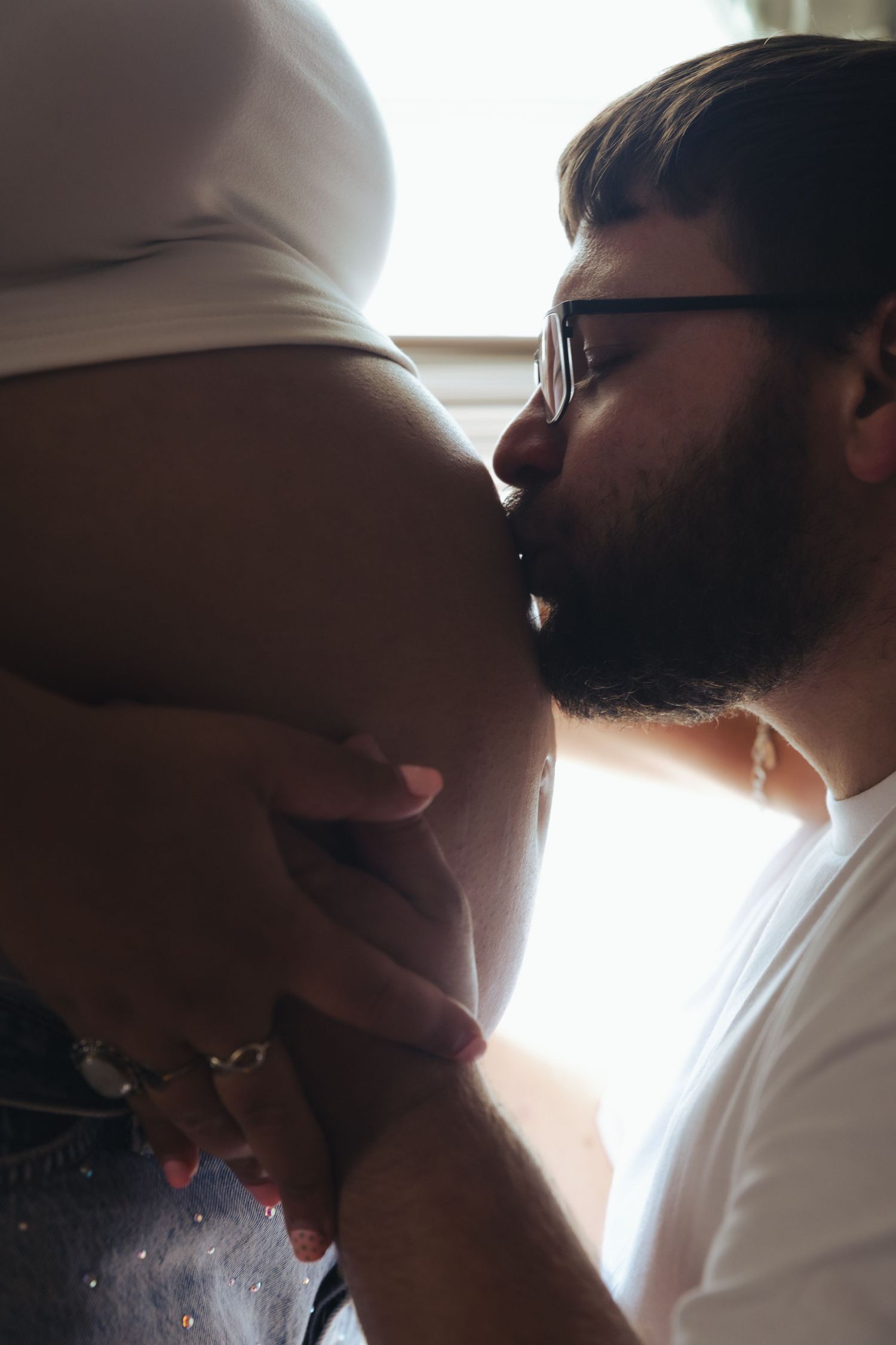 Bearded man with glasses kissing pregnant woman's baby bump, tender maternity moment captured in soft natural light.