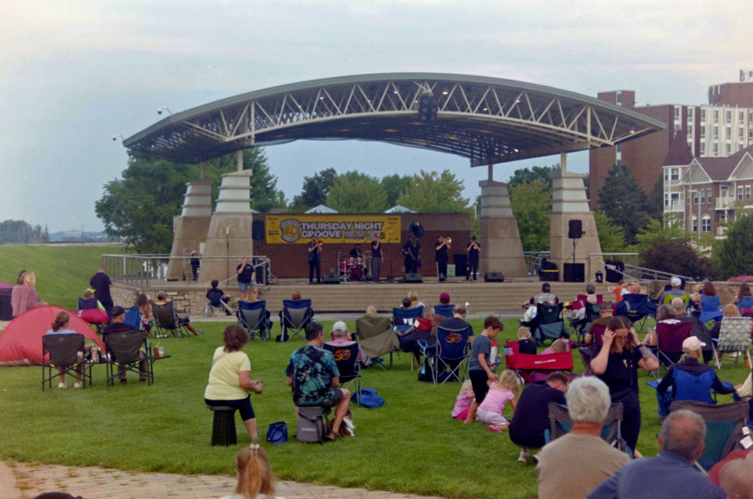 Outdoor concert at a waterfront amphitheater with audience seated on lawn chairs on a cloudy day.