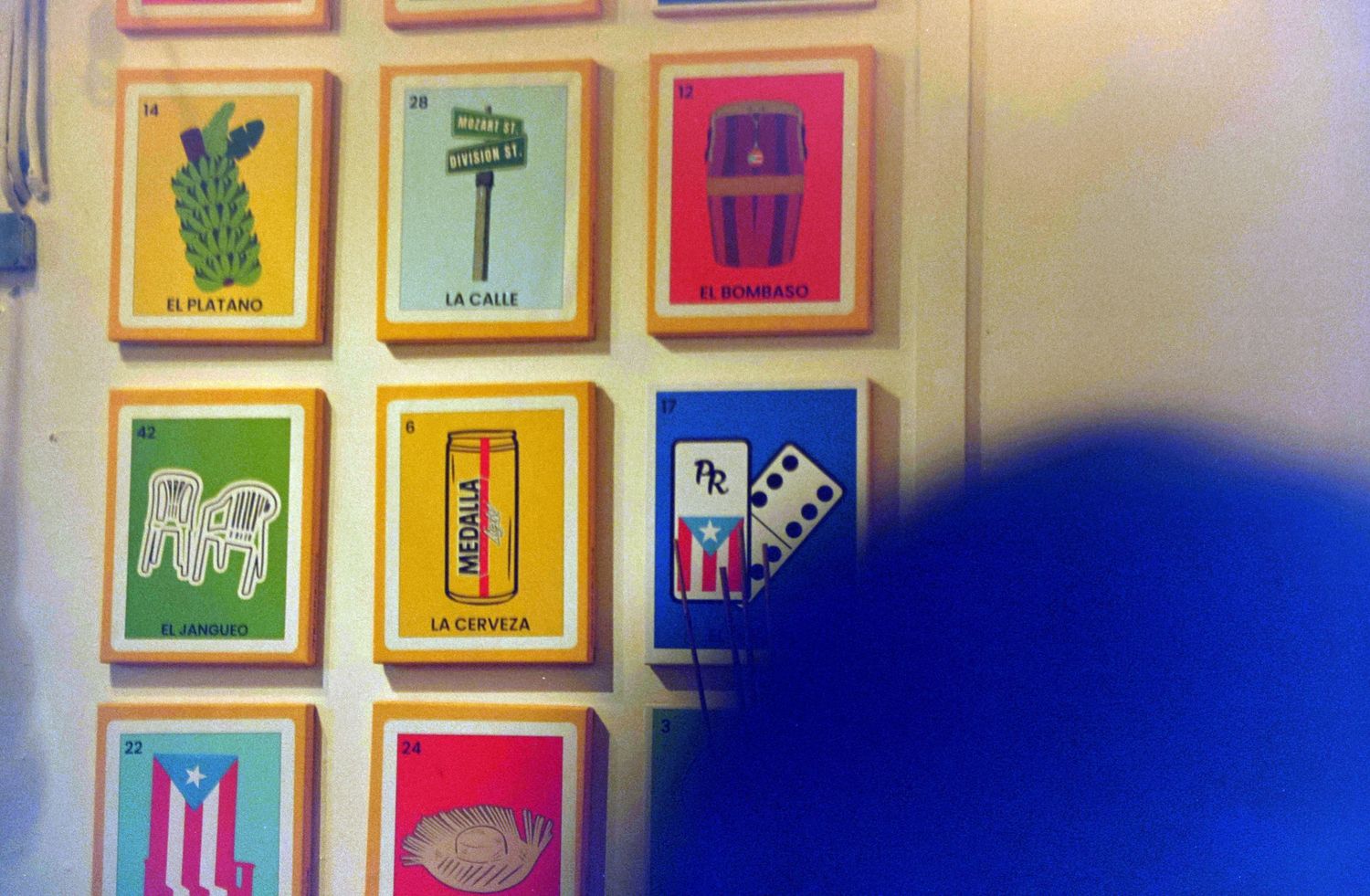 Colorful Mexican loteria cards displayed on a wall featuring illustrations of a plantain, street sign, and dominos.