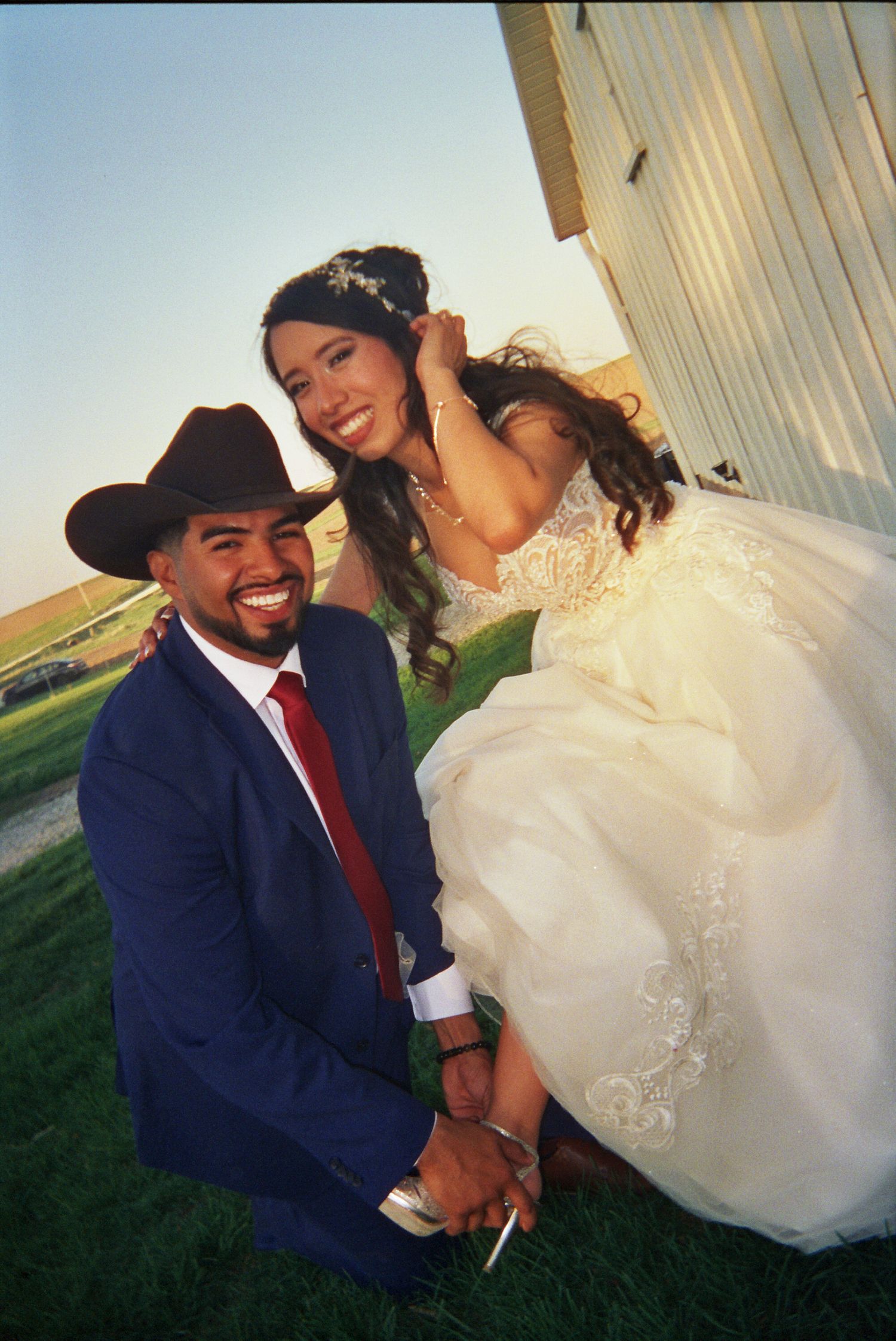 A happy bride in a flowing white dress poses with her groom wearing a navy suit and cowboy hat in a rustic outdoor setting.