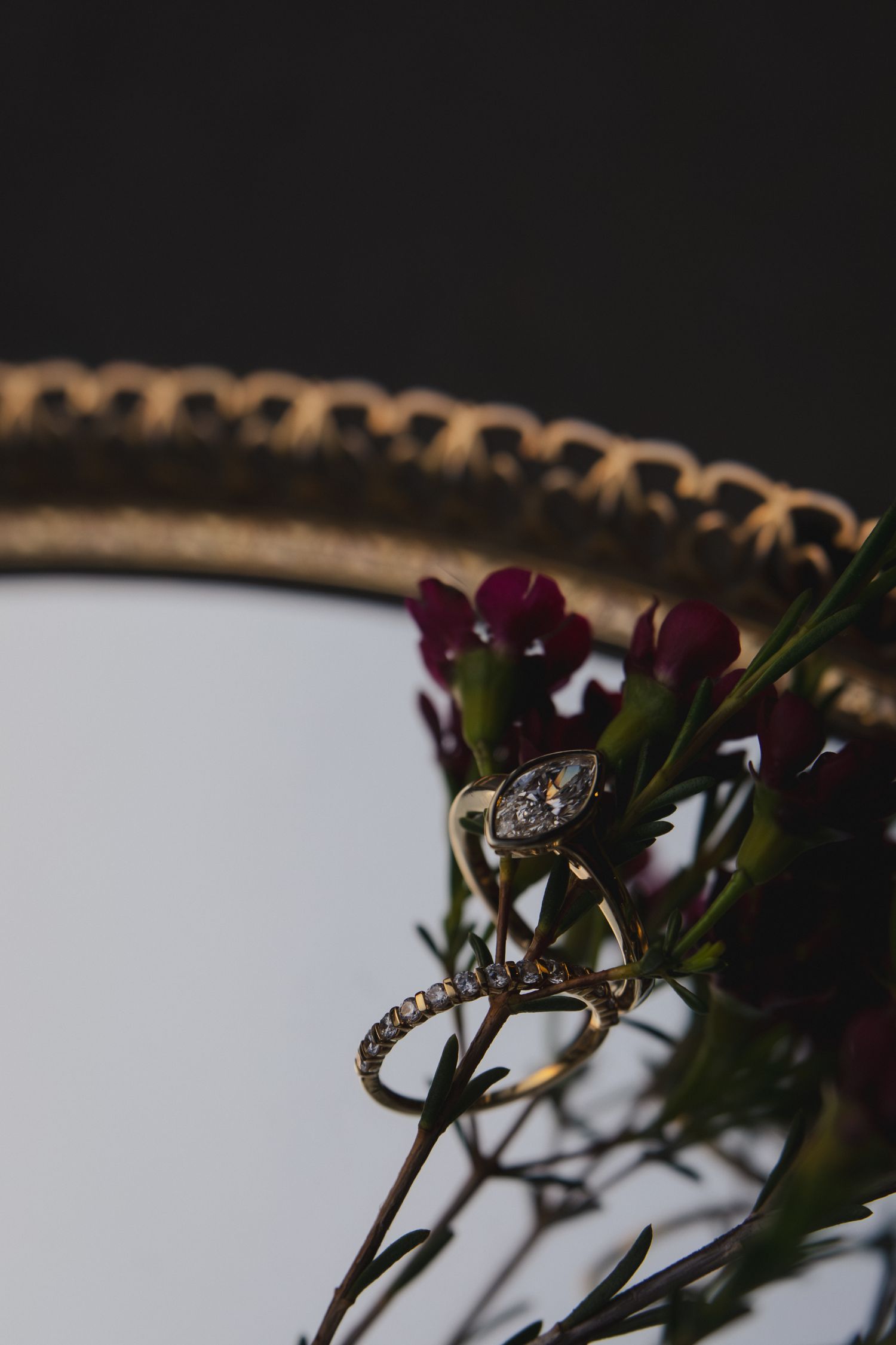 Close-up macro photo of a diamond engagement ring nestled among purple flowers on a golden mirror frame.