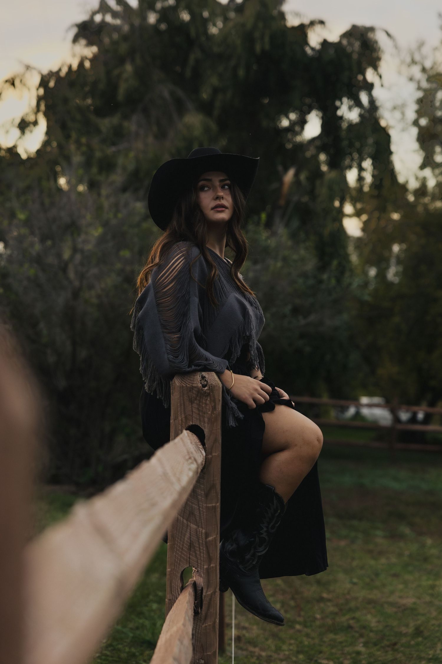 A moody outdoor portrait of someone in a wide-brimmed black hat and dark clothing leaning against a wooden fence at dusk.