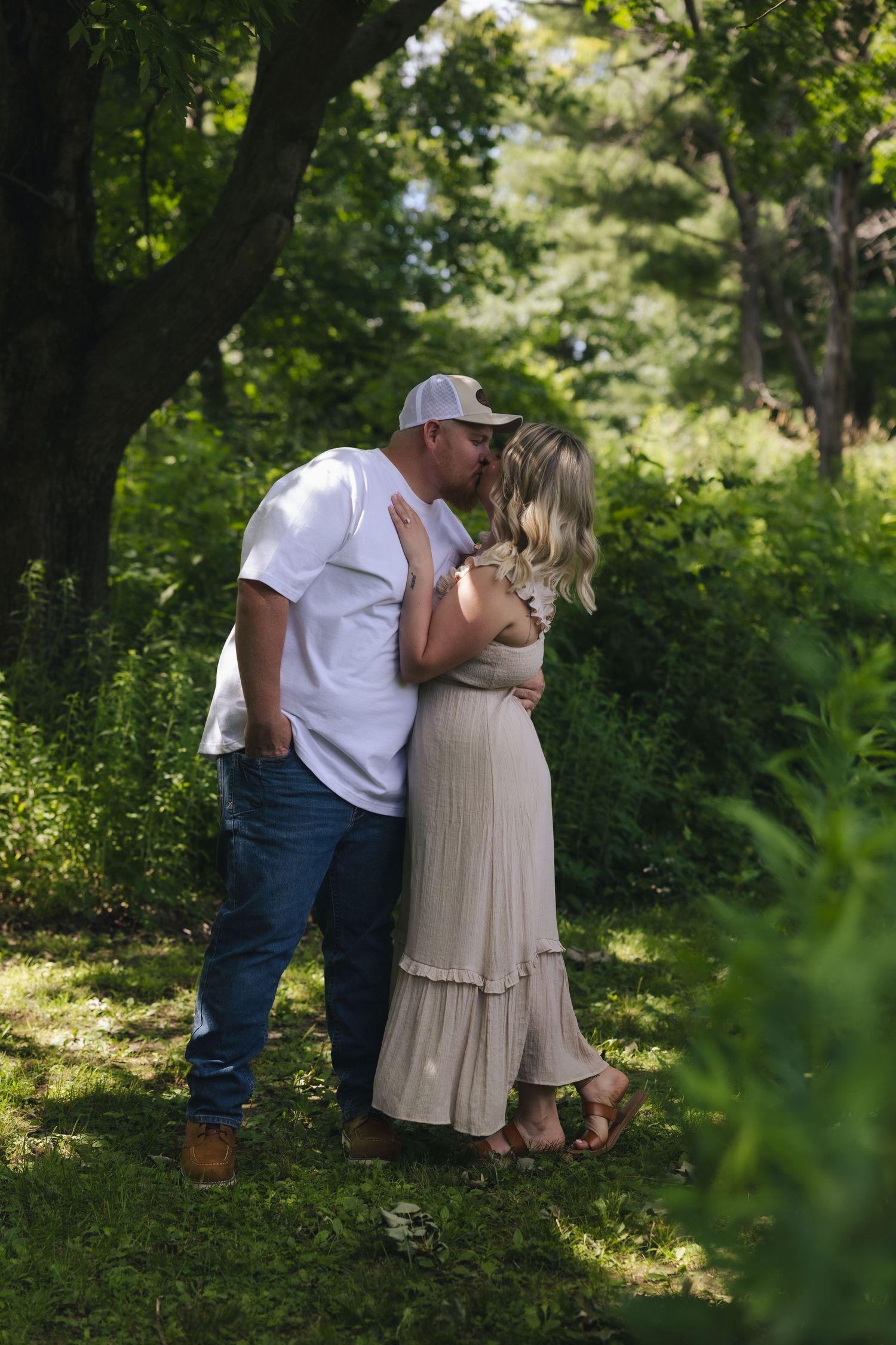 Pregnant couple sharing a kiss outdoors in lush green garden, man in white tee and jeans, woman in beige maxi dress.