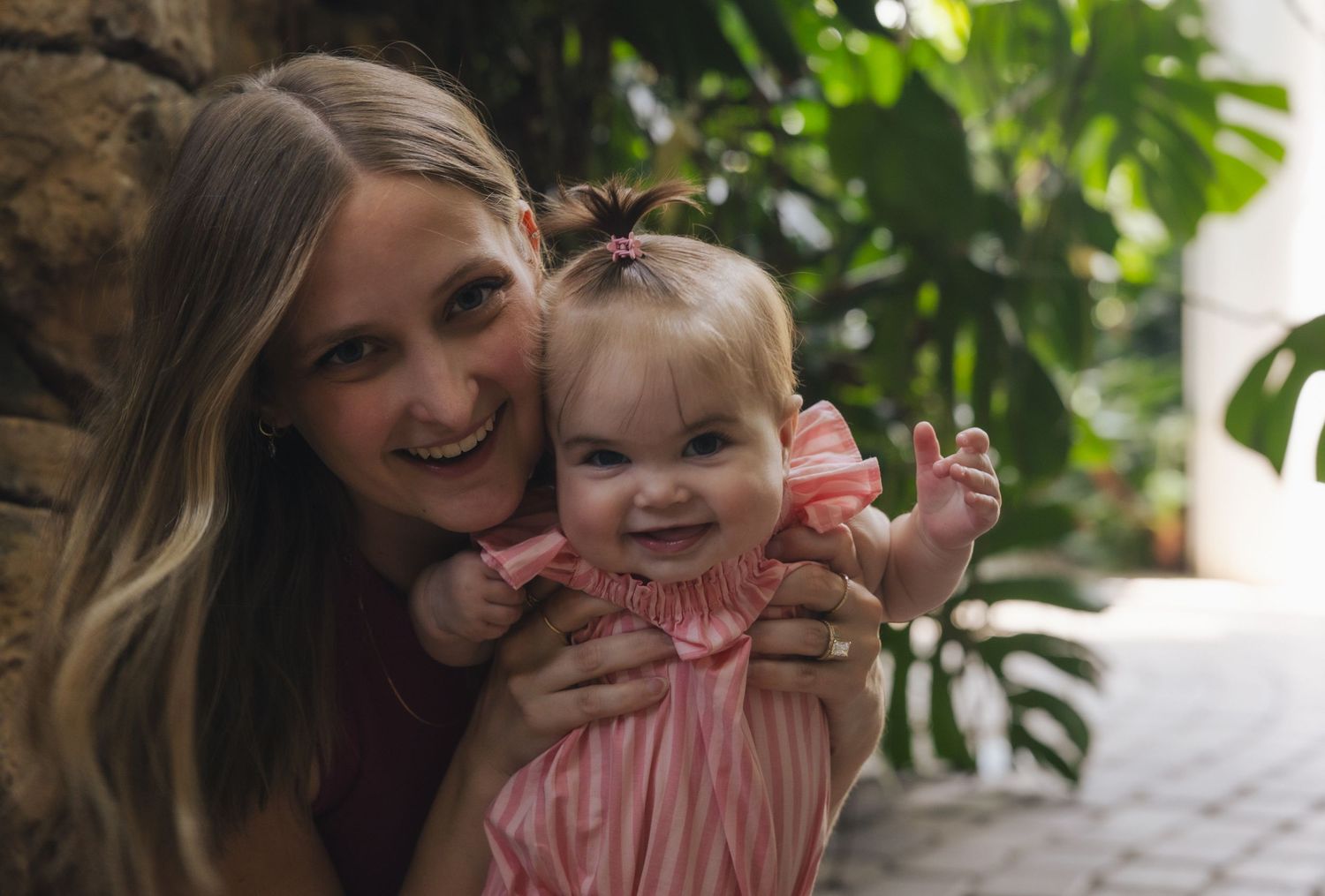 A smiling mother holds her happy baby girl in a pink dress outdoors near lush green tropical plants.