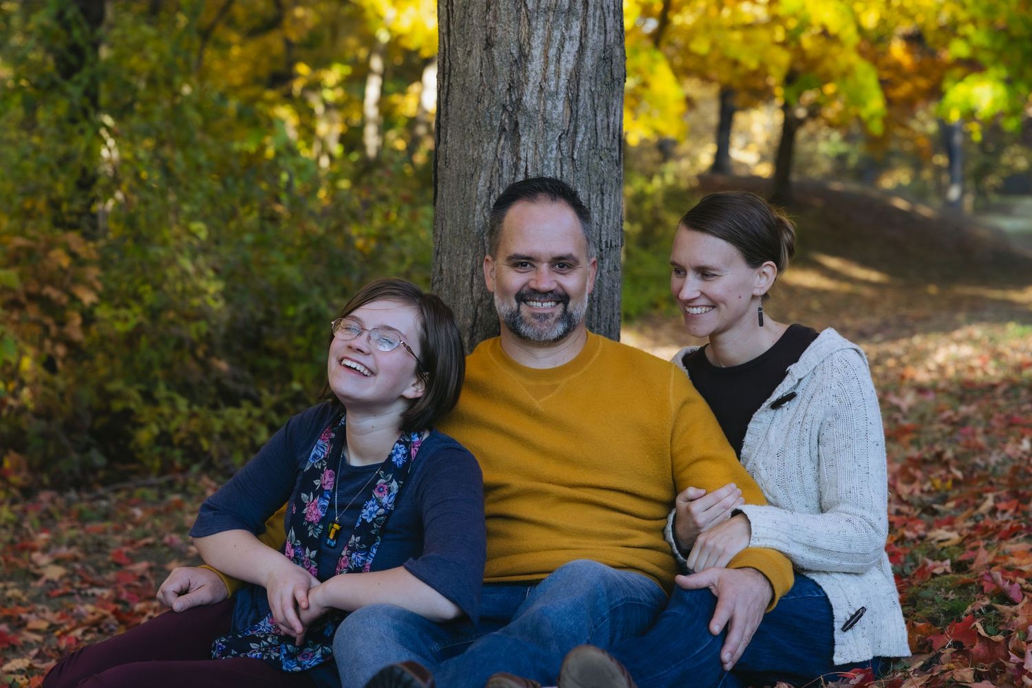 Family of three smiling and laughing together outdoors in autumn park with colorful fall foliage.