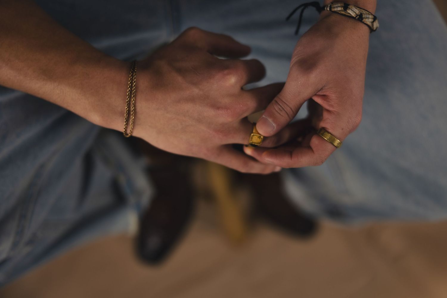 Close-up of hands with gold rings and bracelets clasped together, wearing jeans, with brown boots visible below.