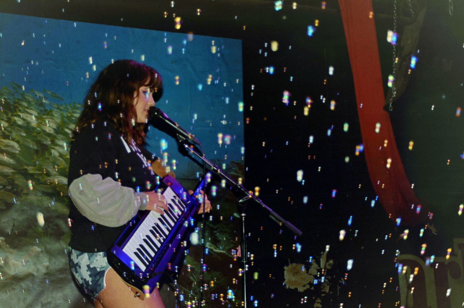 Female musician playing a blue keytar and singing into a microphone on a colorful stage.