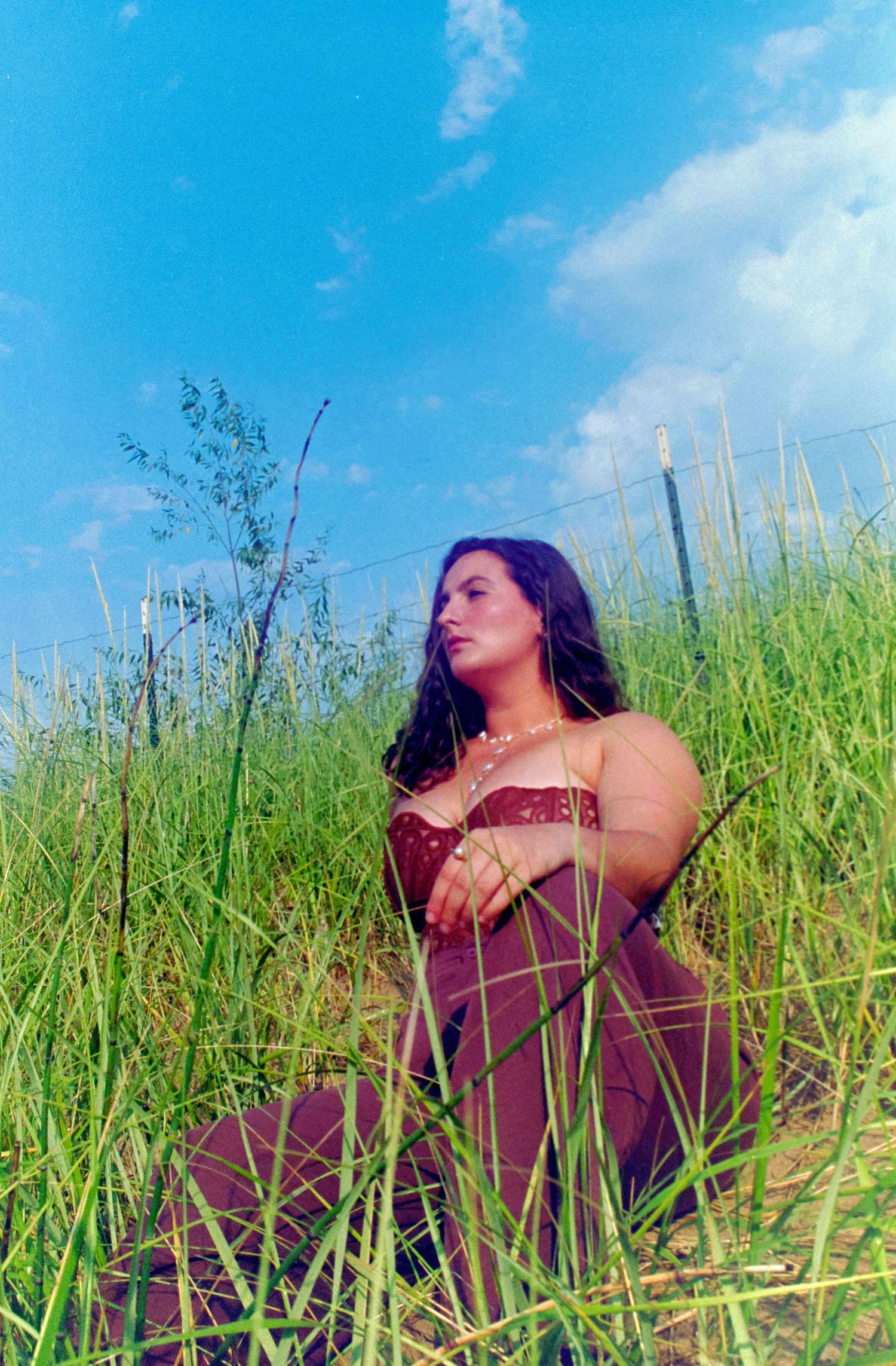 A young woman in a burgundy dress sits peacefully among tall green grass under a bright blue sky.