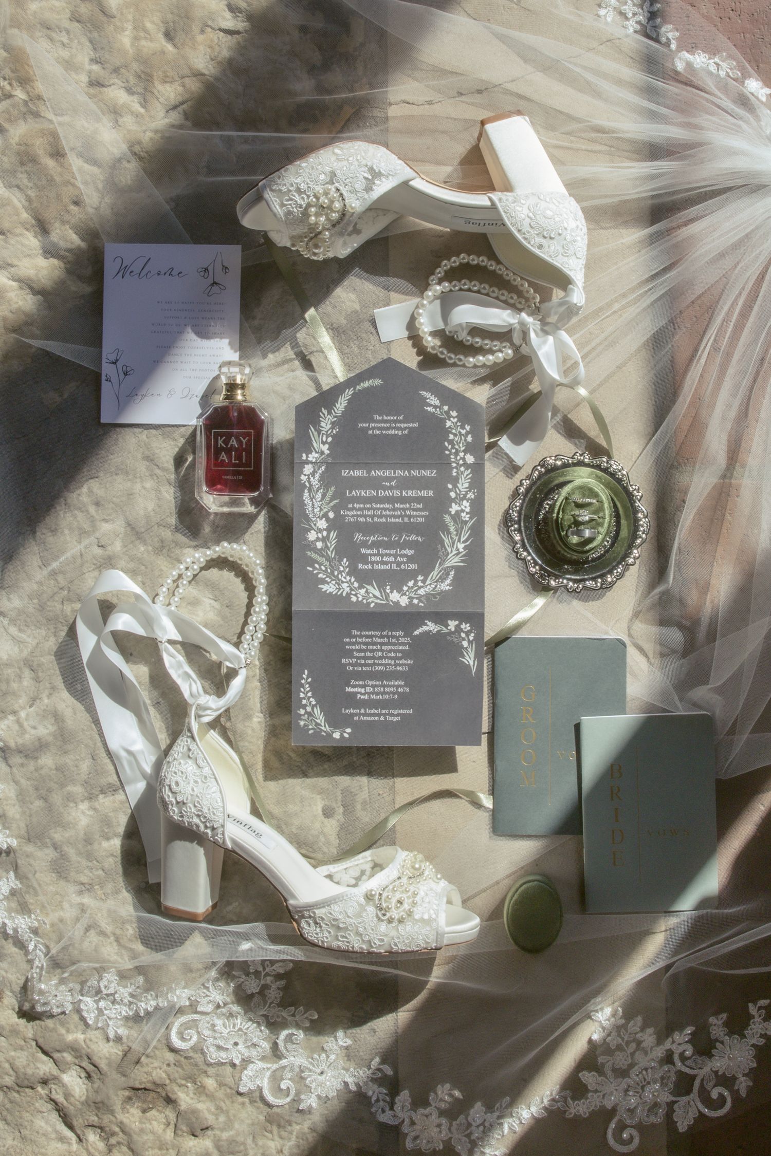 Wedding details flatlay with lace shoes, invitations, perfume bottles and veil on a textured neutral background.