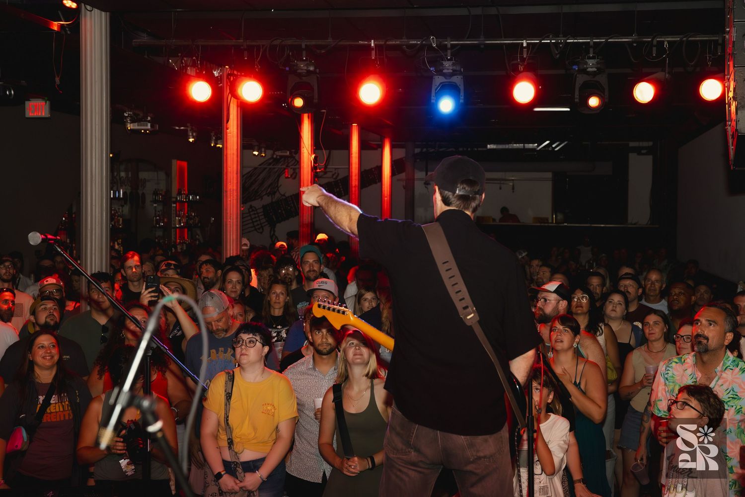 Crowded indoor concert venue with colorful red and blue stage lighting illuminating a packed audience on the dance floor.