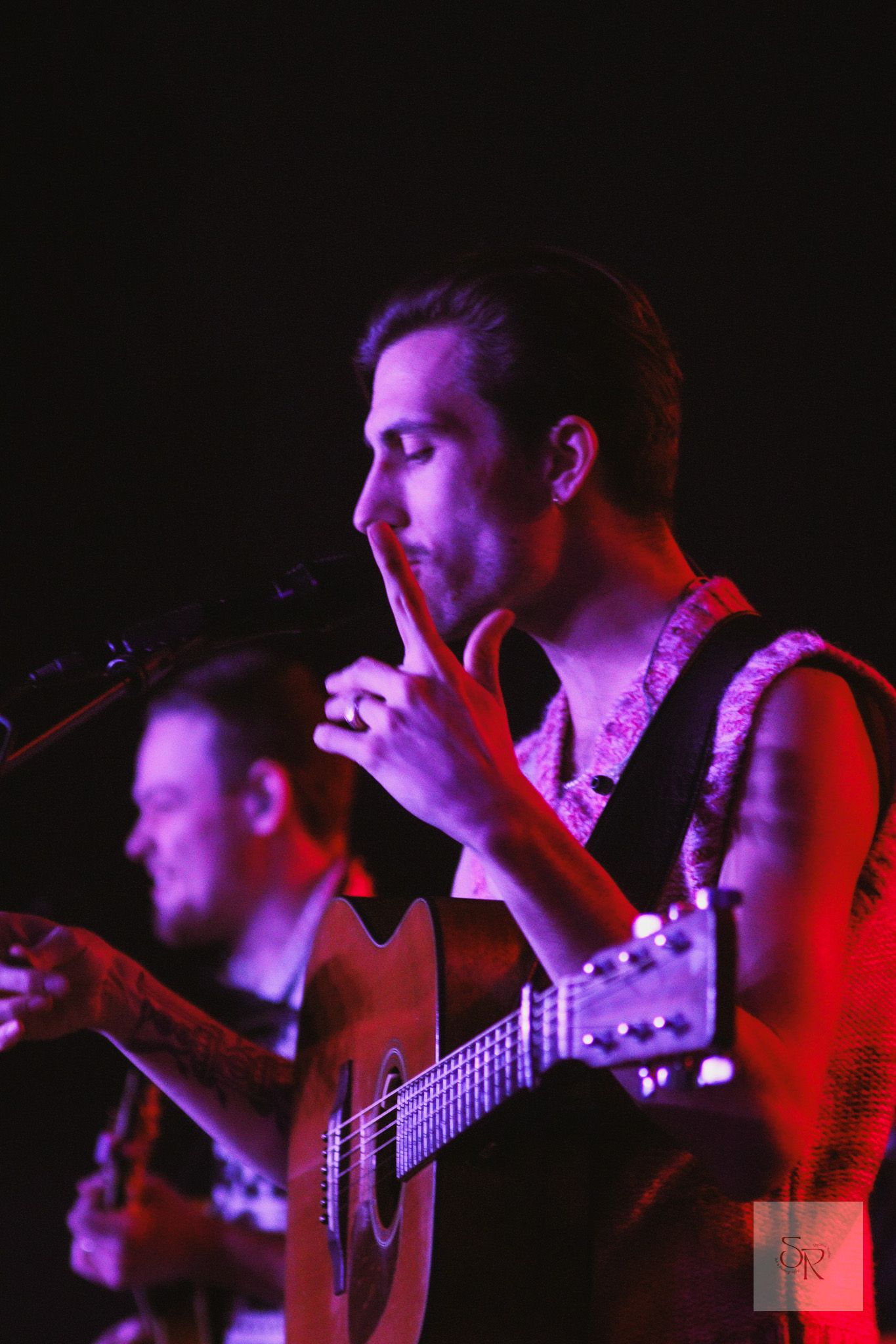 Silhouetted musicians performing on stage with acoustic guitars illuminated by dramatic red and purple lighting.