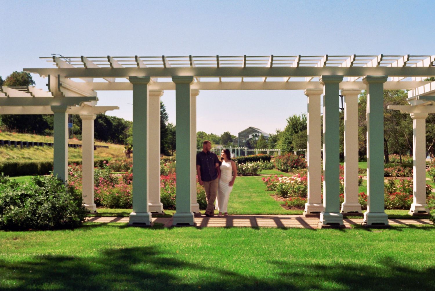 A white pergola structure sits atop manicured grass with decorative gardens visible at sunset.
