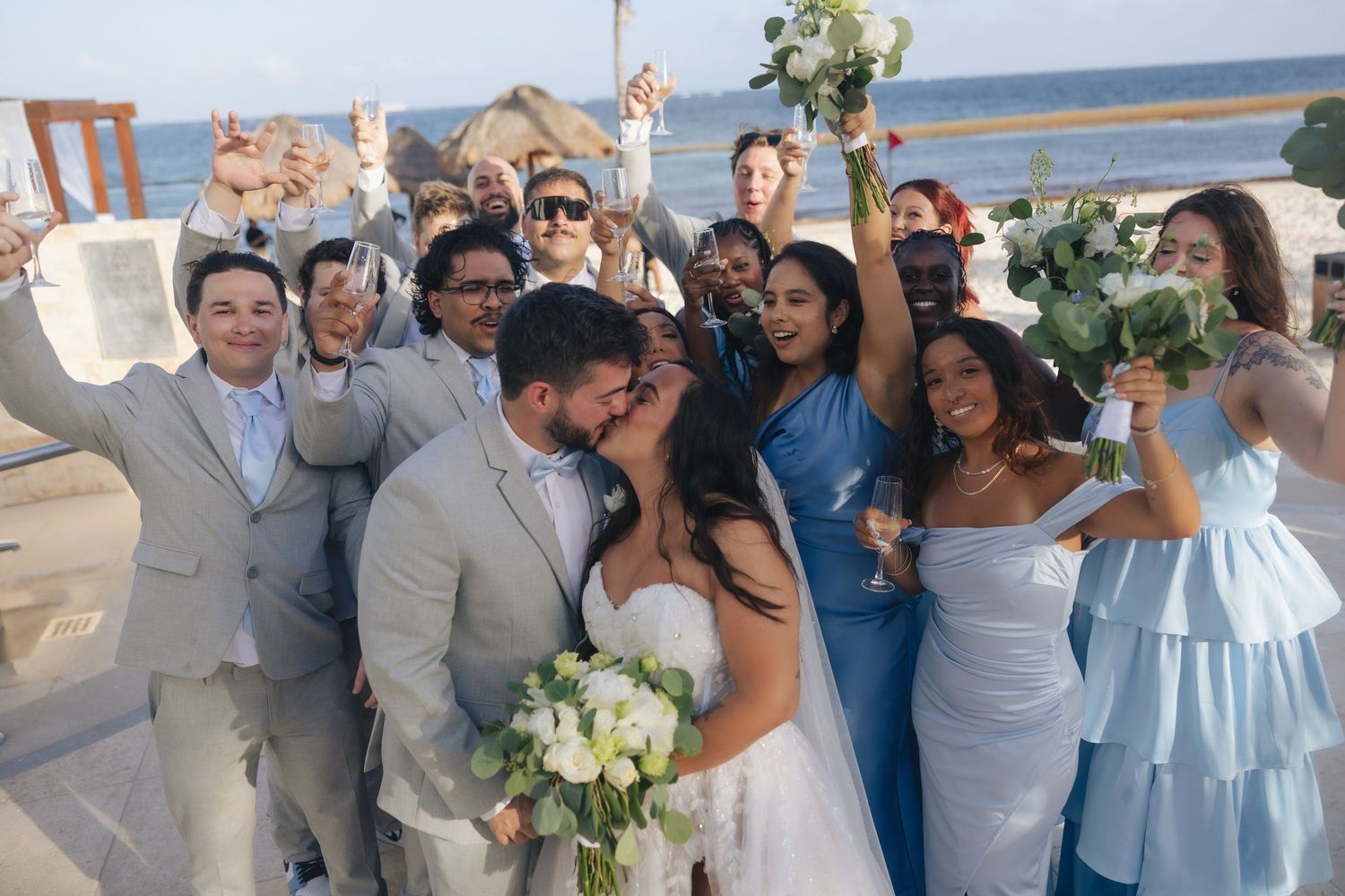Bride and groom kiss surrounded by cheering wedding party in blue attire with ocean backdrop.