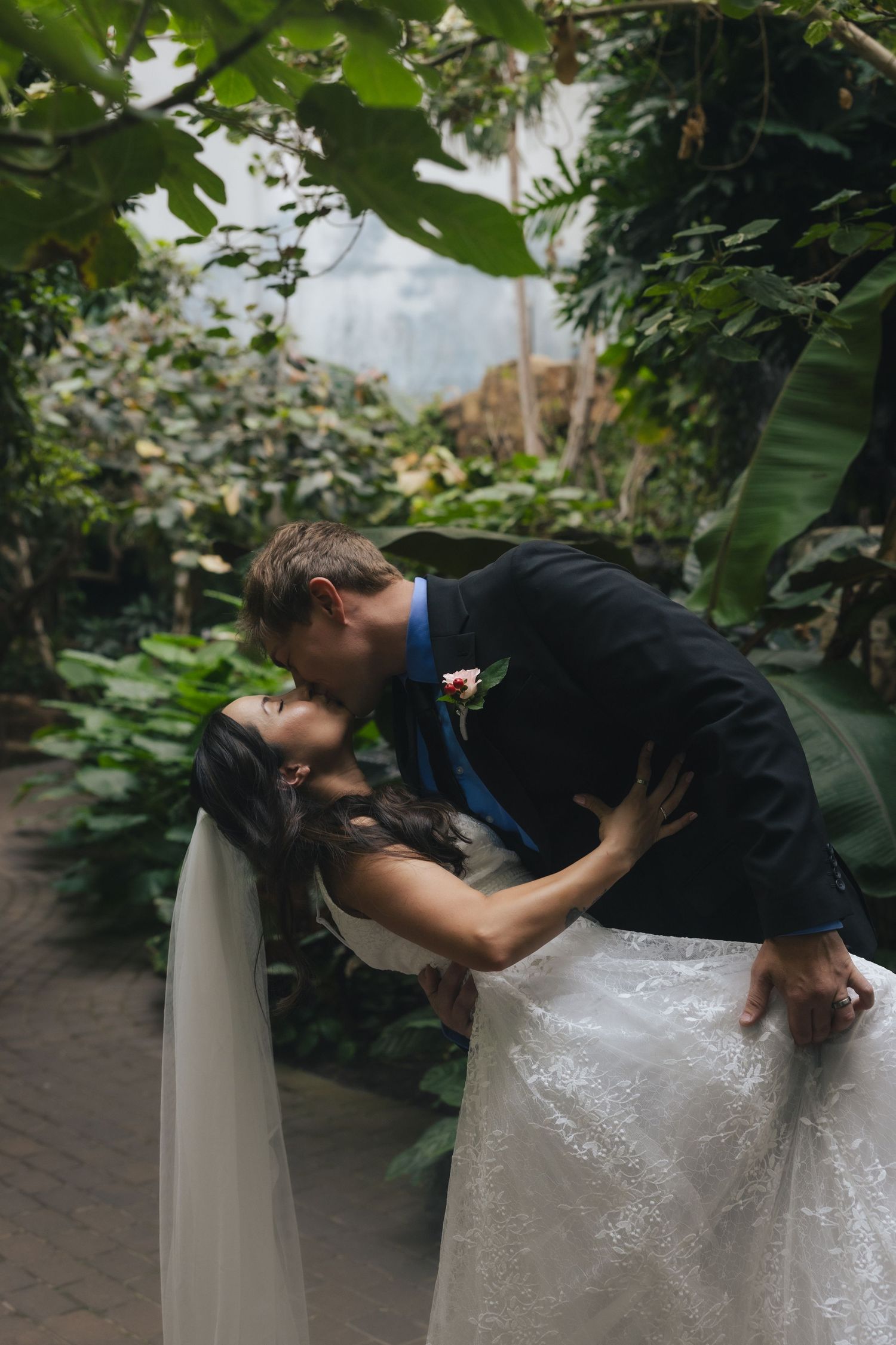 Bride and groom sharing a romantic dip kiss surrounded by lush tropical greenery in an indoor botanical garden.