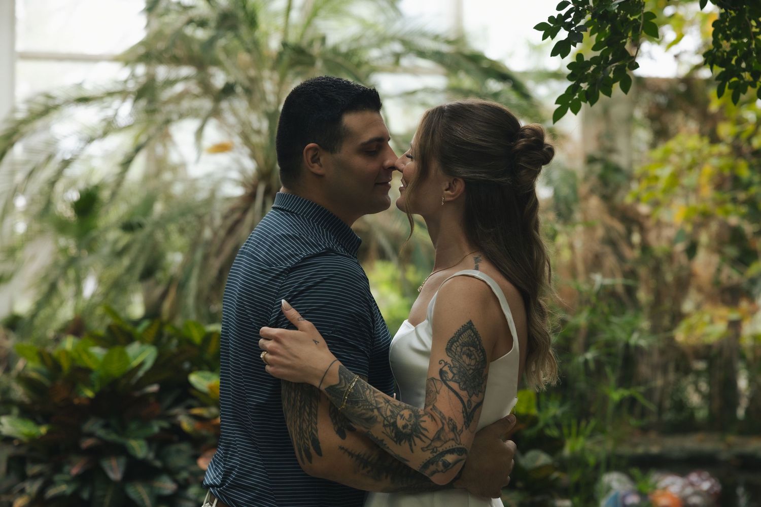 Couple sharing a romantic moment in a lush greenhouse, man with tattoos embracing woman in white dress.