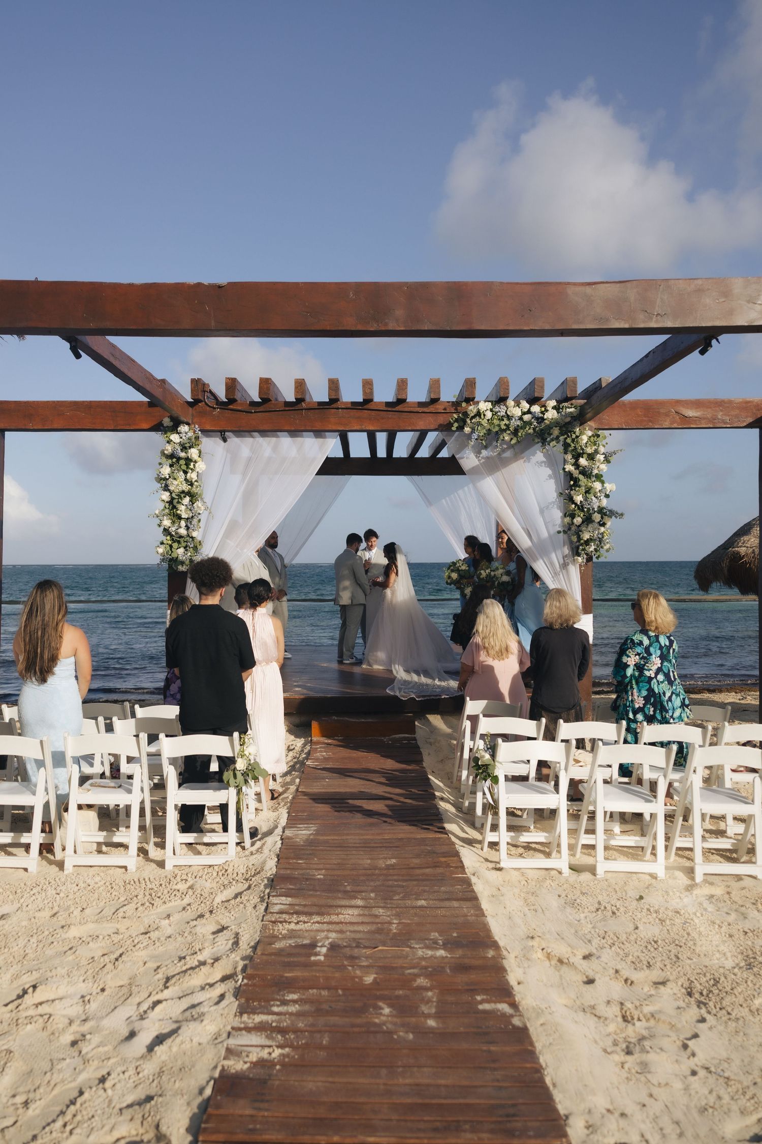 Beach wedding ceremony under a wooden pergola with white drapes and floral decor, ocean backdrop, guests seated on white chairs.