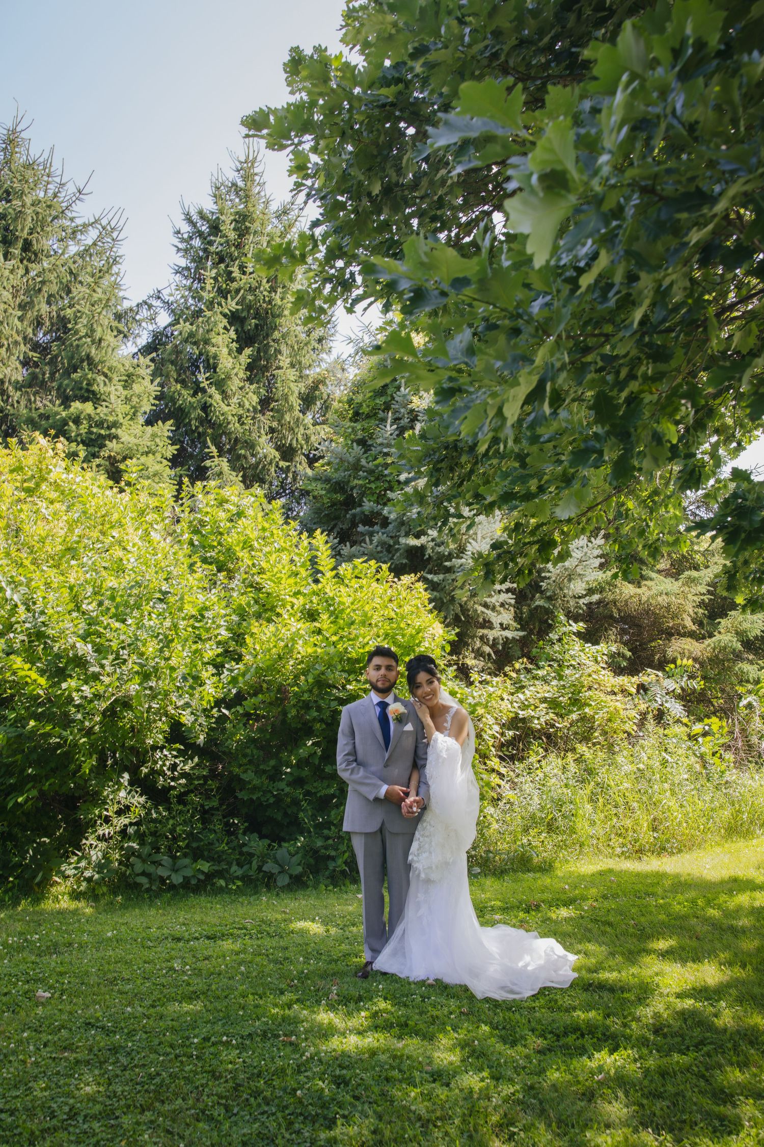 A couple poses together on a grassy hill surrounded by lush green trees and foliage on a bright summer day.