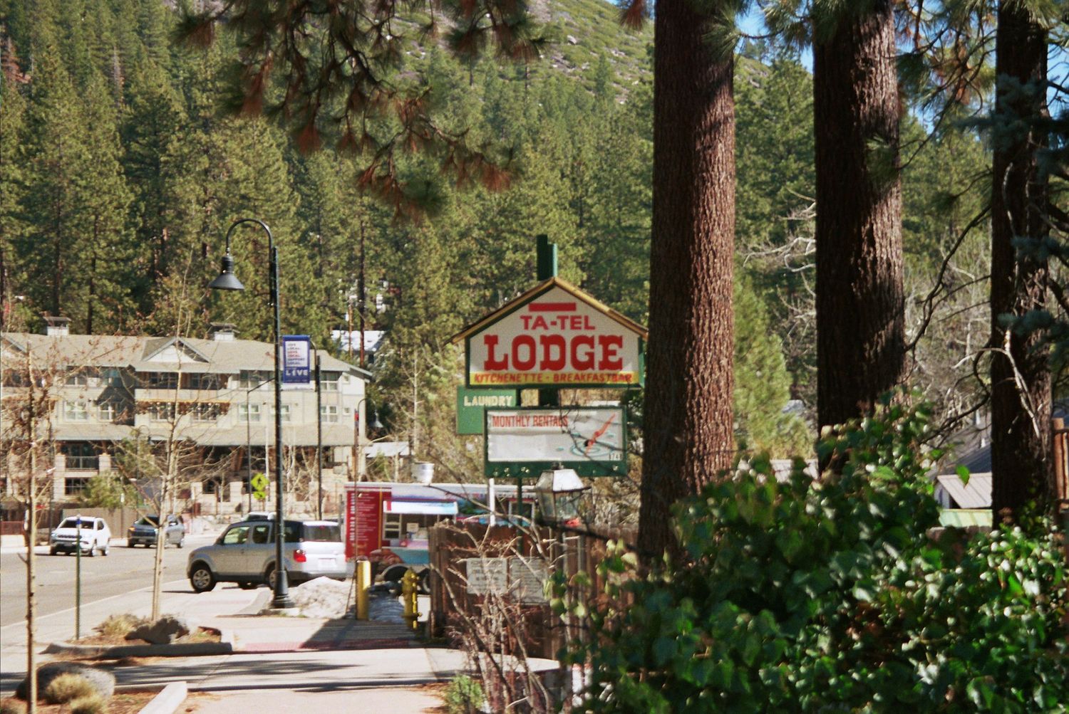 Travel Lodge sign surrounded by tall pine trees in a mountain resort town with parked cars and shops nearby.
