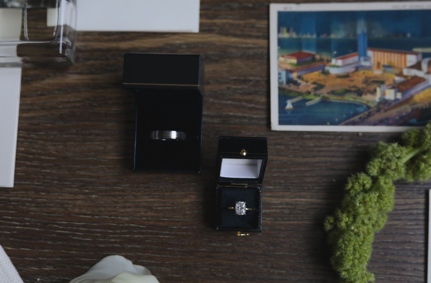 Two elegant diamond rings displayed in open black jewelry boxes on a wooden table with a colorful cityscape photo nearby.