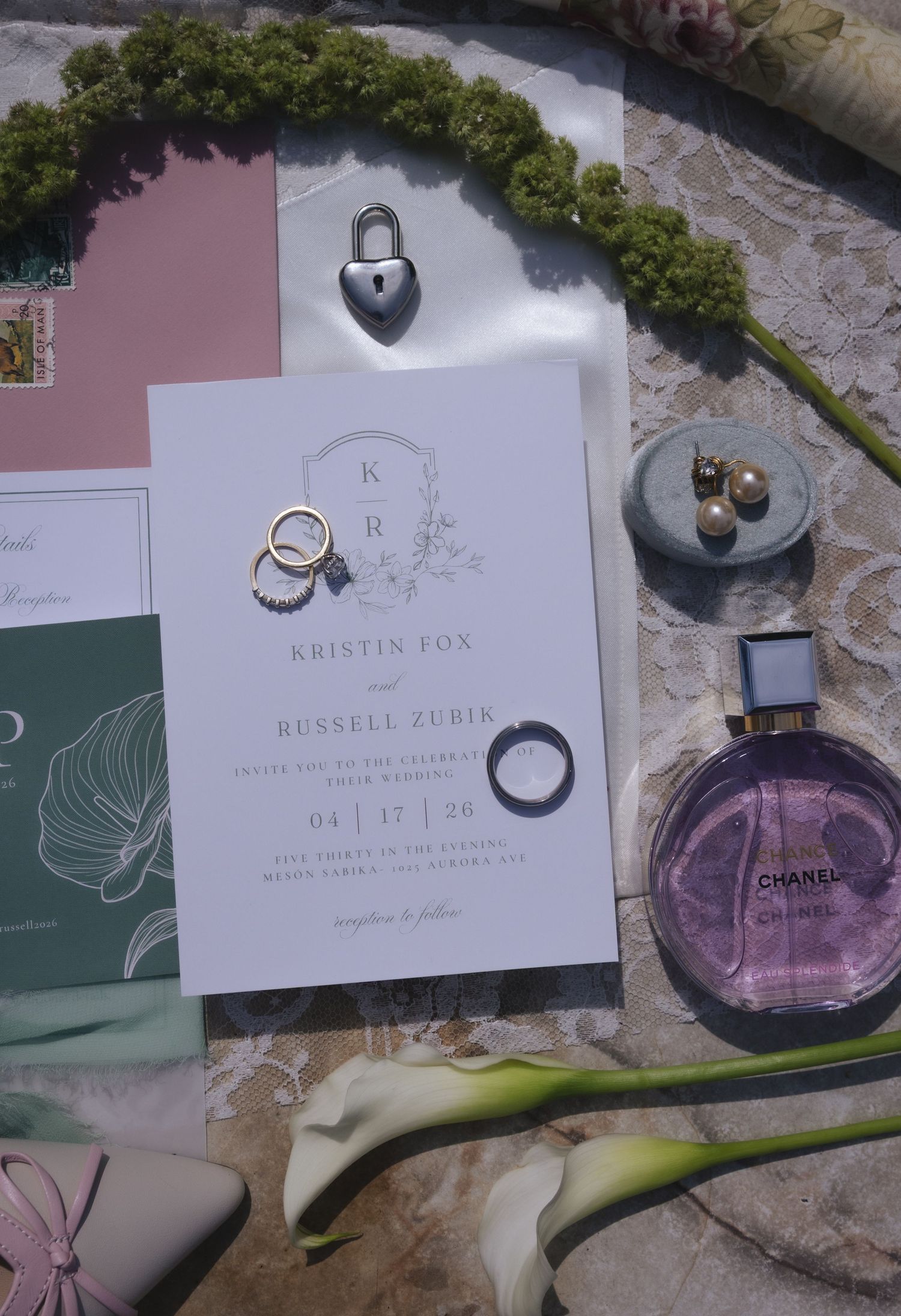 Elegant wedding flat lay featuring Kristin Fox and Russell Zubin invitation, gold rings, Chanel perfume, heart lock, and floral accents.