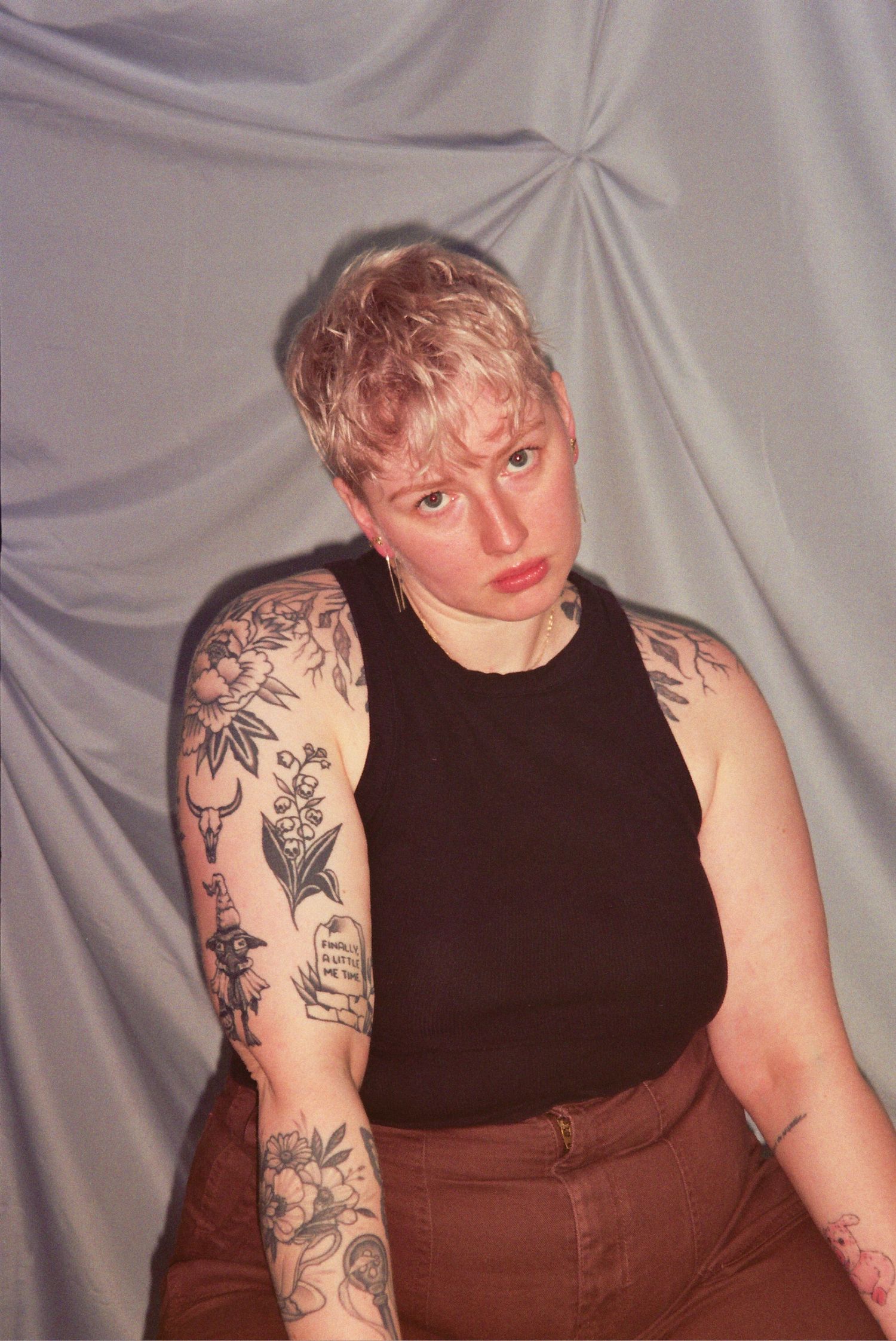 Person with short blonde hair and tattoos wearing a black tank top and brown pants posed against gray backdrop.