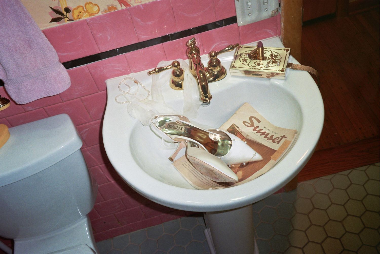 White pedestal sink with gold faucet, a high heel shoe, and Sunset magazine in a pink tiled bathroom.
