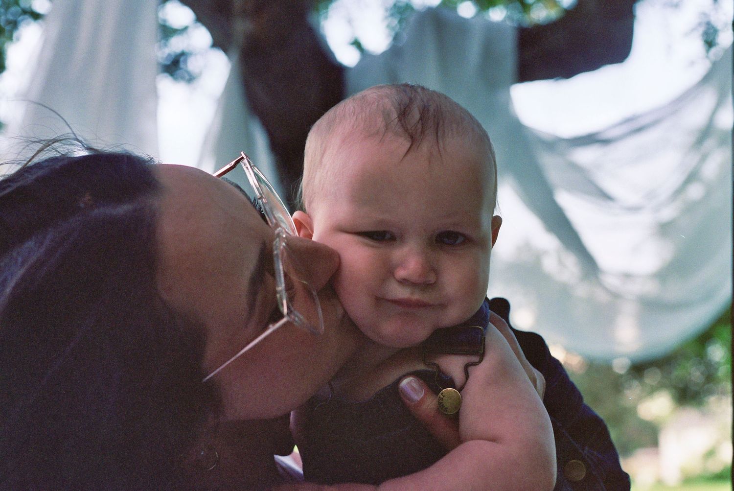 A small baby smiles while being held and receiving a kiss outdoors on a sunny day.