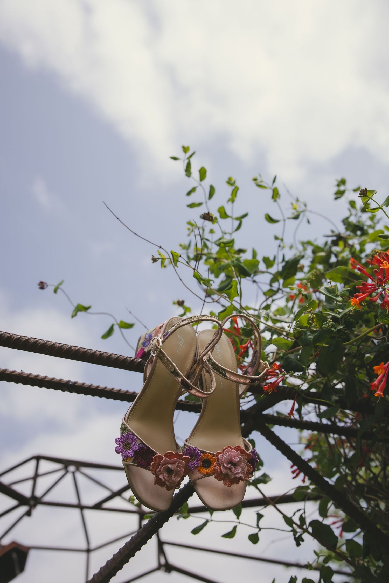 A pair of floral embellished sandals hanging on metal bars against a cloudy sky with green foliage and red flowers.