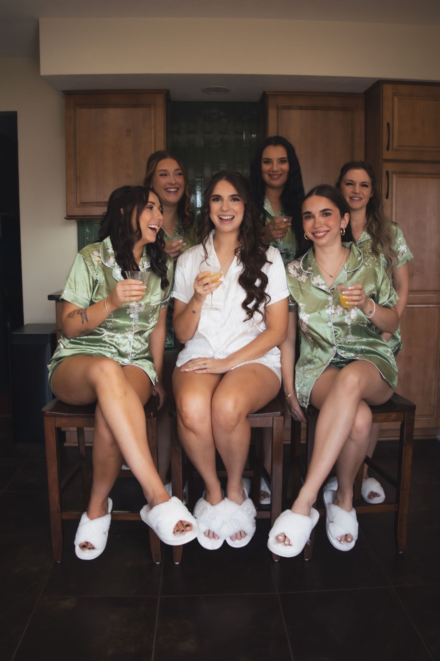 Bride and bridesmaids wearing matching mint green silk robes and white slippers sit together during wedding preparations.