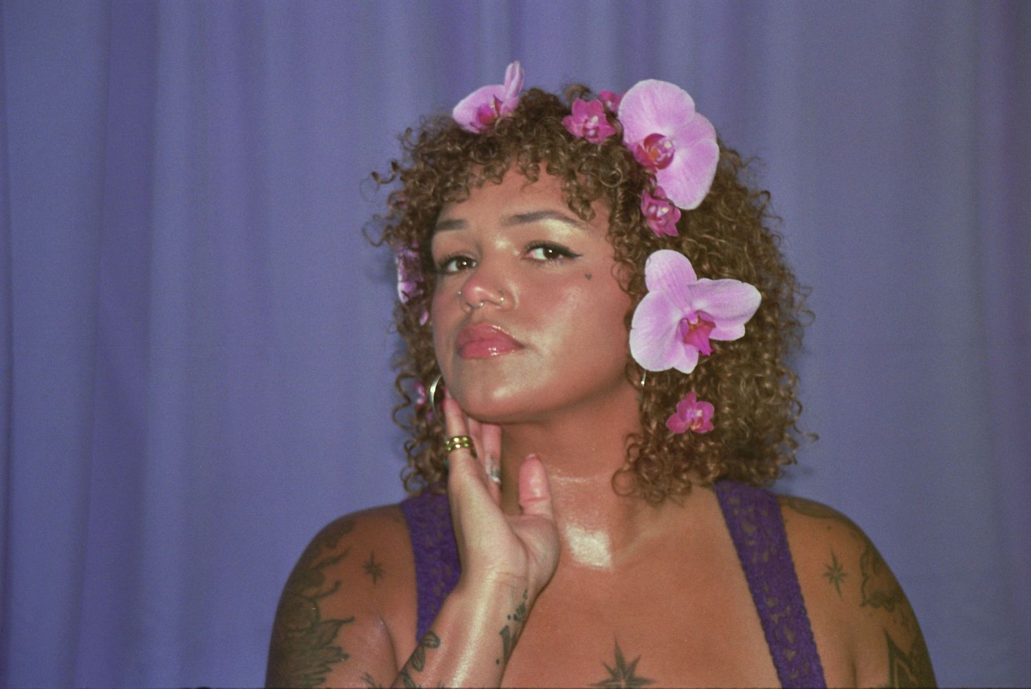 Woman with curly hair adorned with pink orchid flowers, tattoos, and purple top posing against a lavender background.