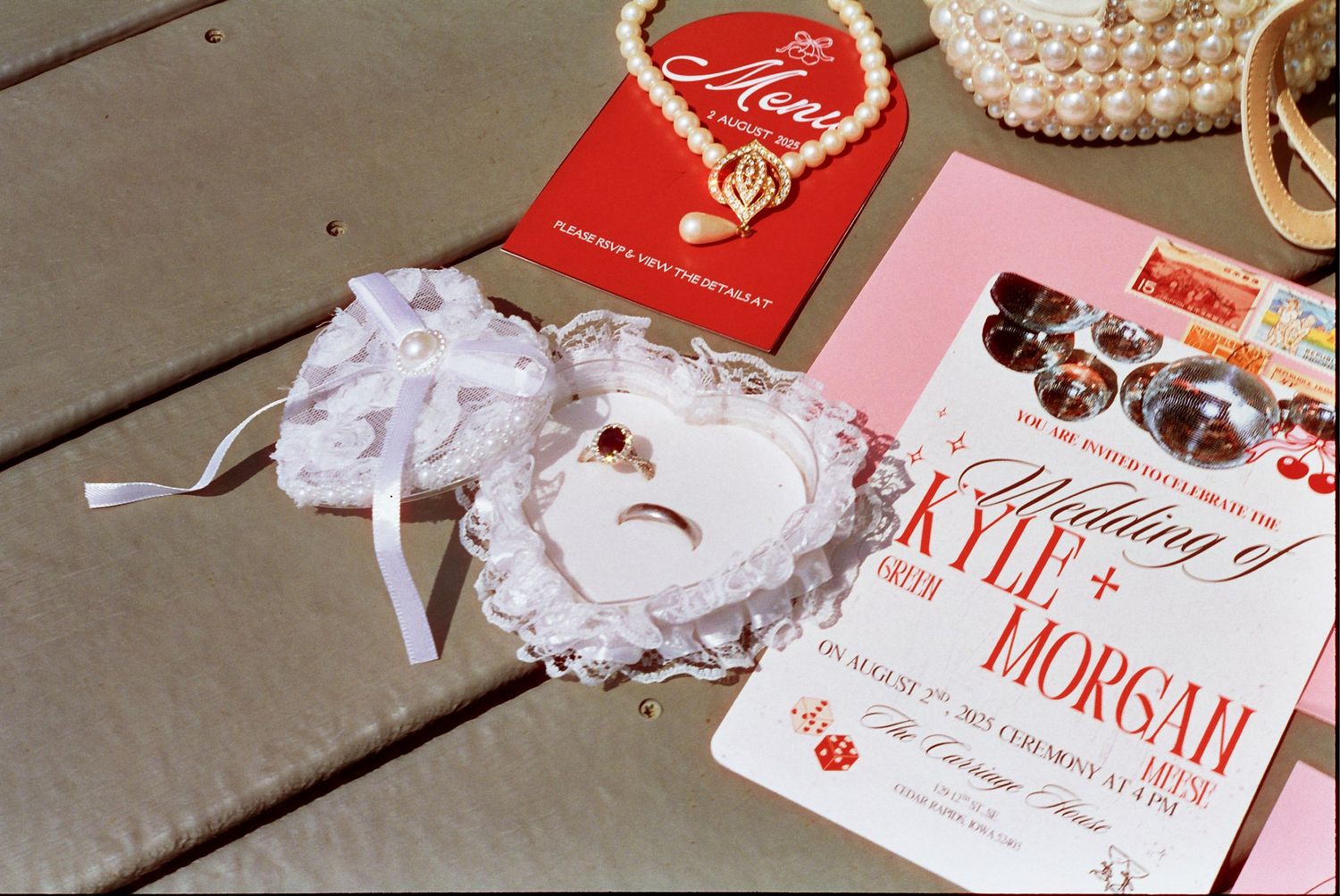 Vintage wedding accessories including white lace garters, a red invitation, and a romantic novel on a wooden surface.