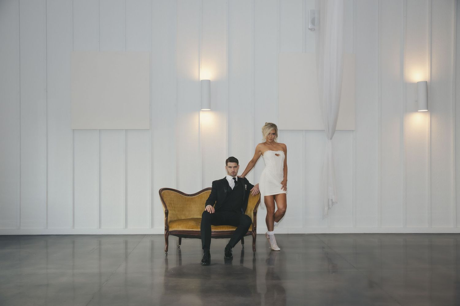 Two people pose in a minimalist white room with wall sconces and a vintage armchair.