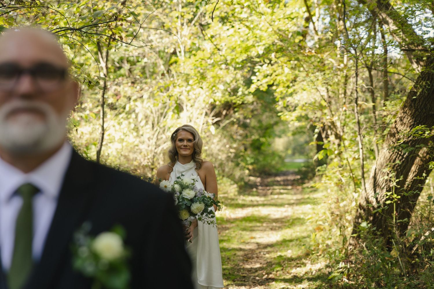A joyful outdoor wedding ceremony takes place on a sunlit wooded path surrounded by green trees and natural scenery.