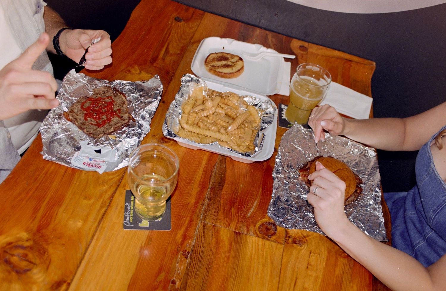 People sharing fast food meal with fries and drinks on foil trays at a wooden table.