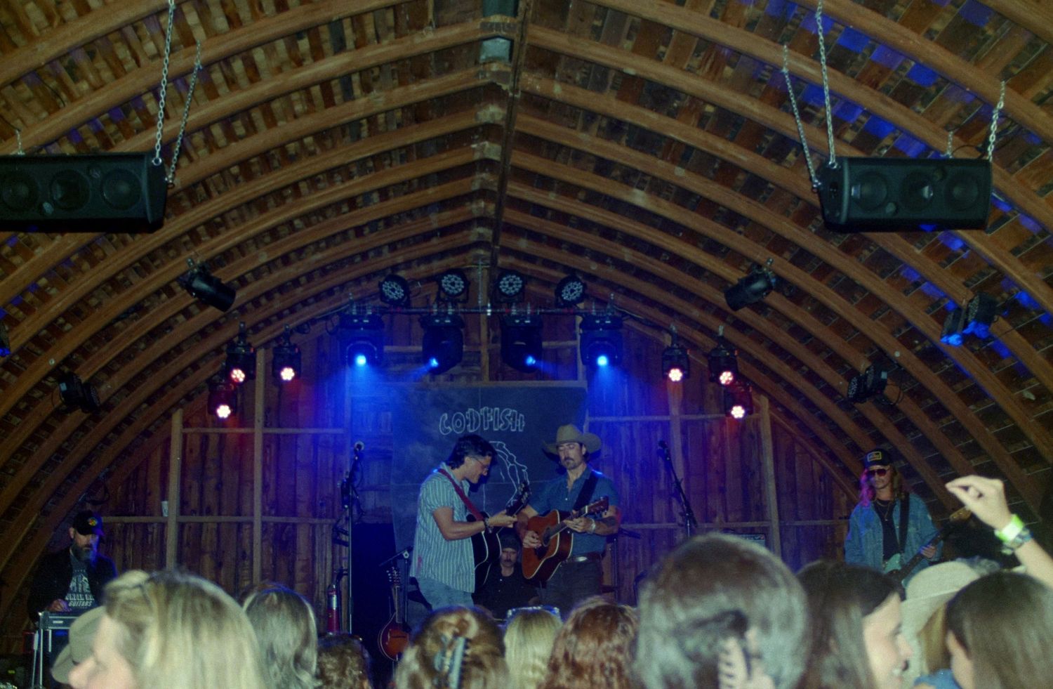 Live band performing on a rustic wooden barn stage with colorful lights and an engaged crowd.