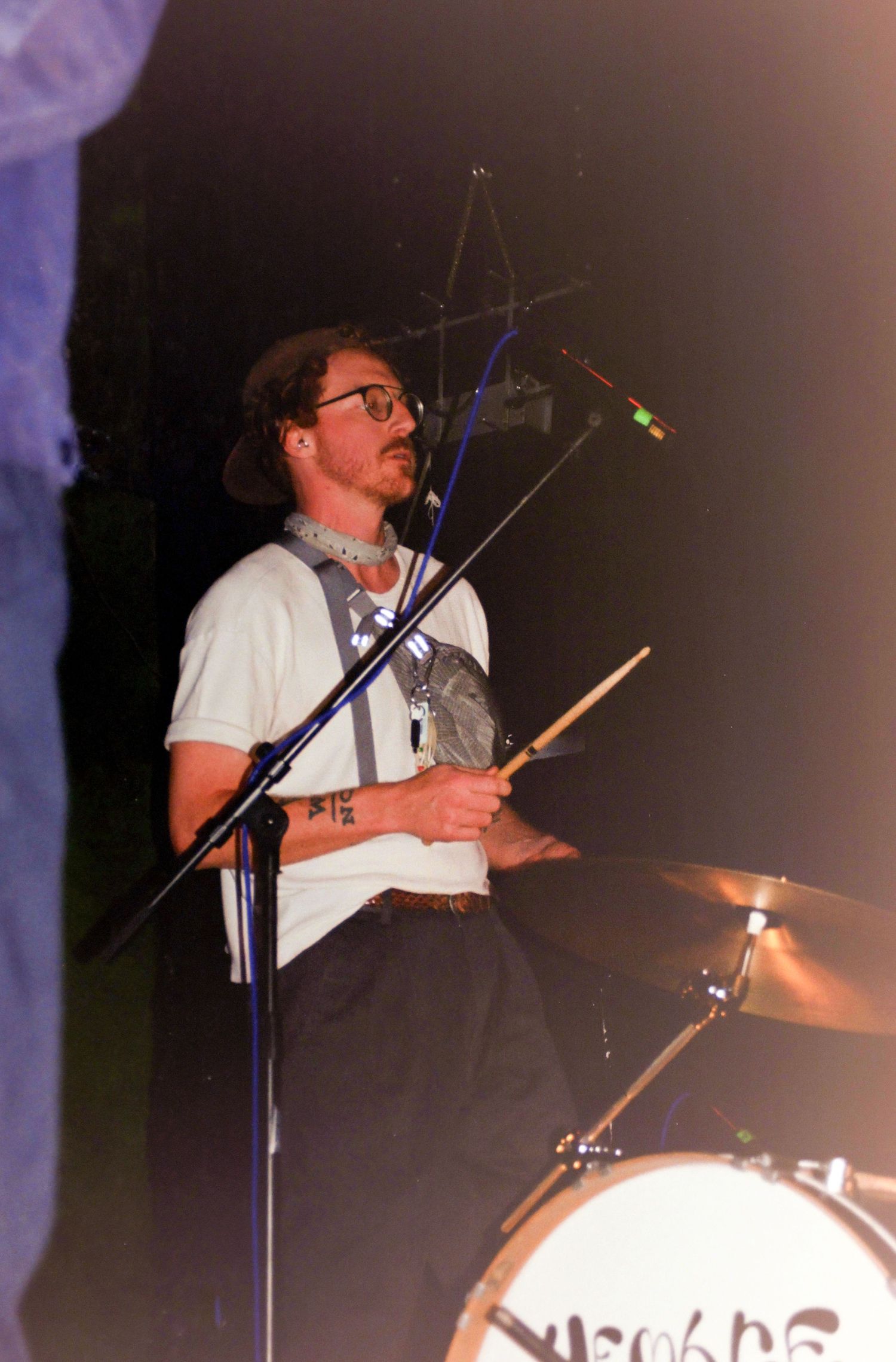 A drummer with glasses and suspenders plays on stage, holding drumsticks near a cymbal and microphone stand.