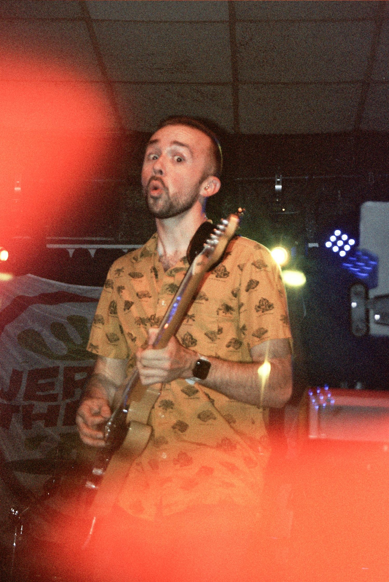 Male guitarist in yellow patterned shirt performs on stage under red and yellow lights at an indoor music venue.