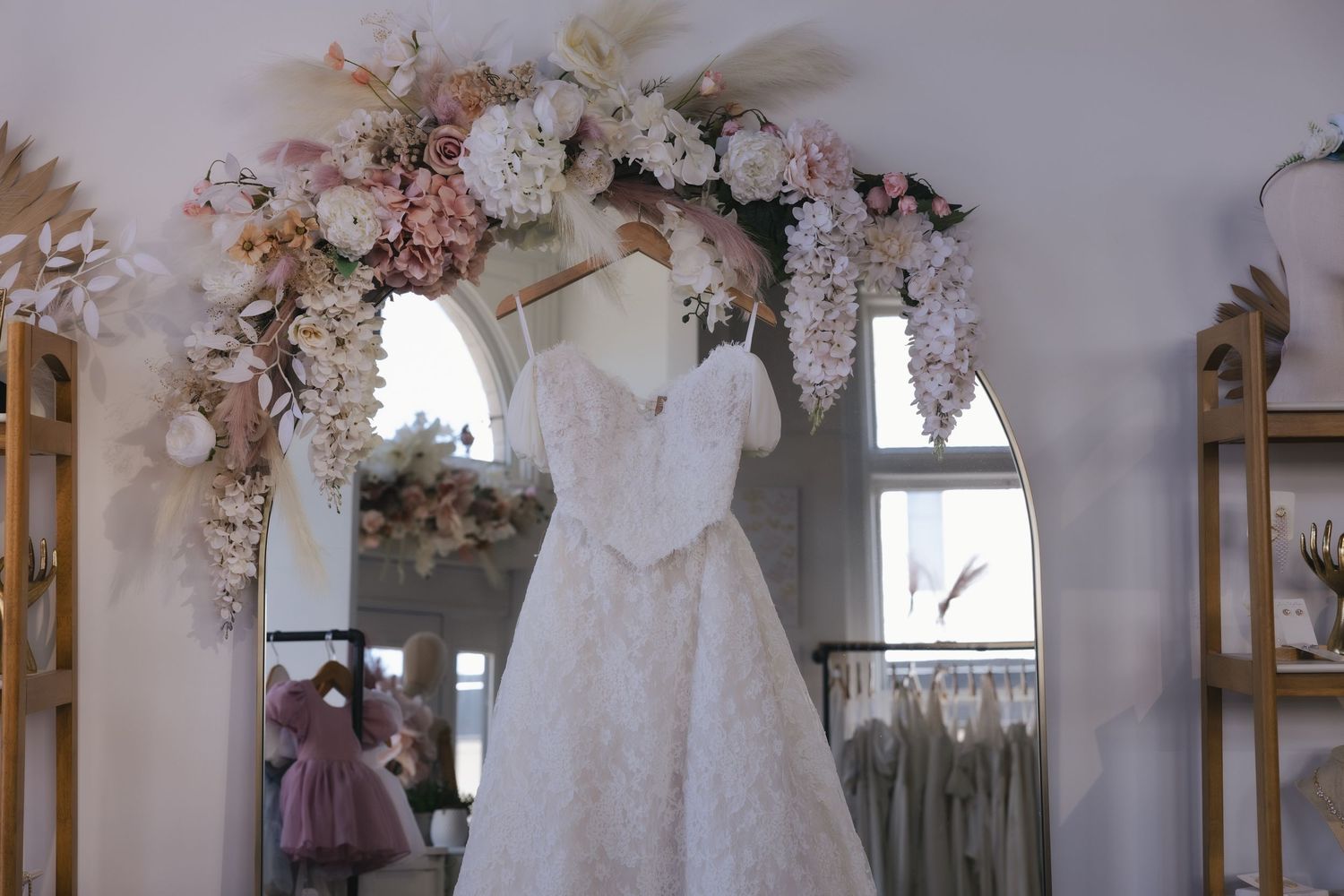 Elegant white lace wedding dress displayed on a hanger beneath a floral arch in a bridal boutique.