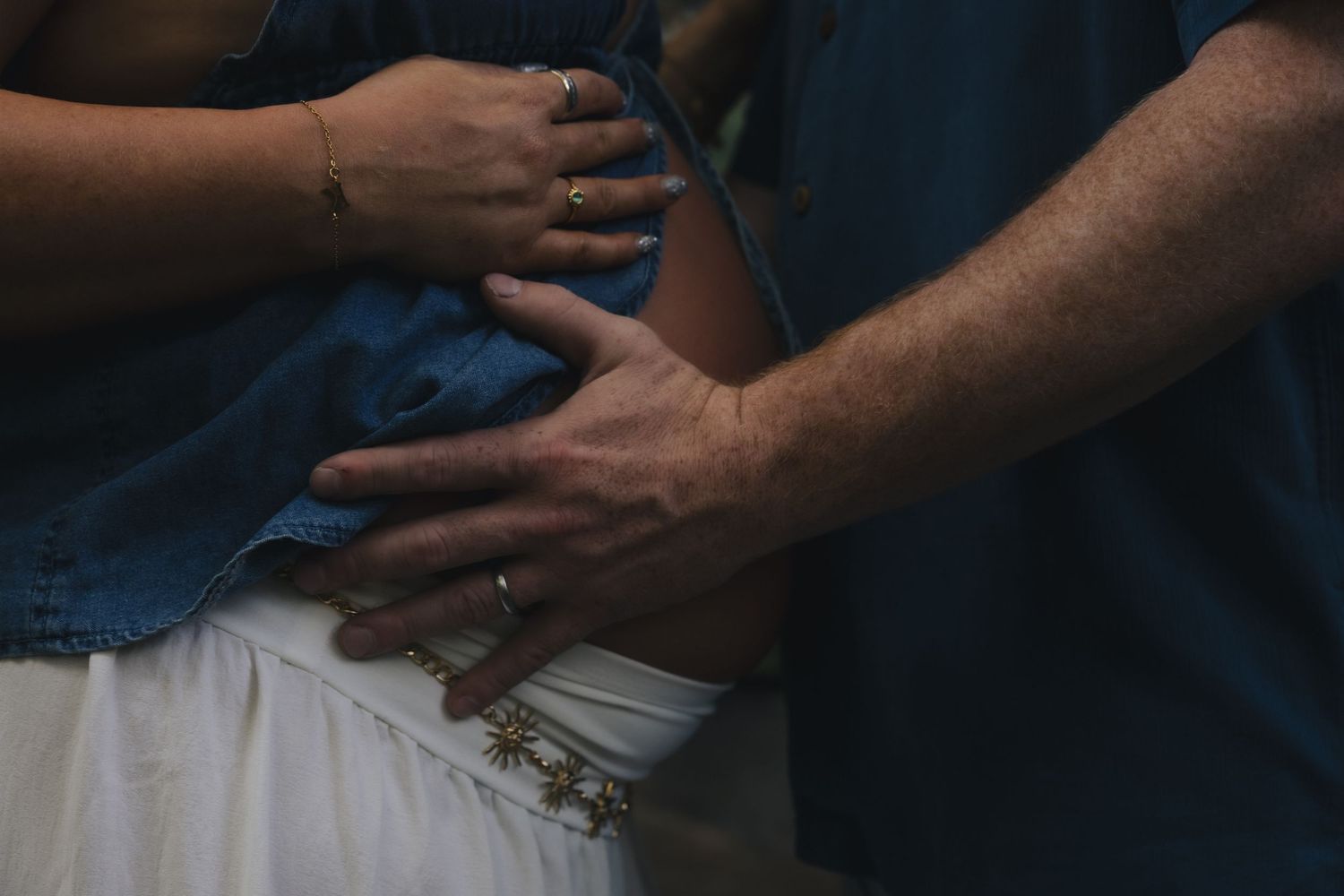 Couple tenderly holding hands on a pregnant belly, wearing rings and bracelets, in a soft outdoor maternity photo.