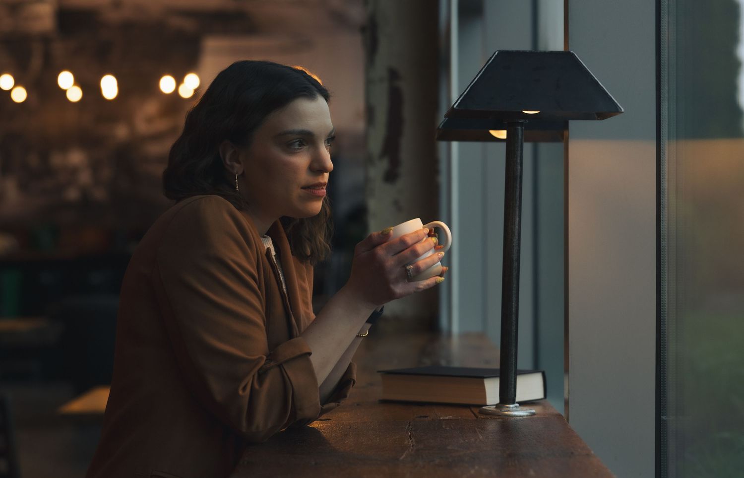 Woman holding coffee mug gazes thoughtfully out window beside modern table lamp in cozy cafe setting.