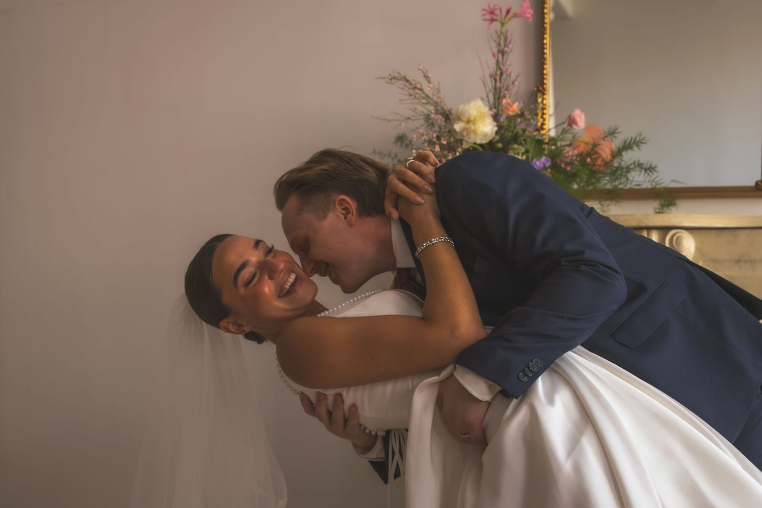 A couple in formal attire embrace and laugh together during a romantic moment at an indoor venue.