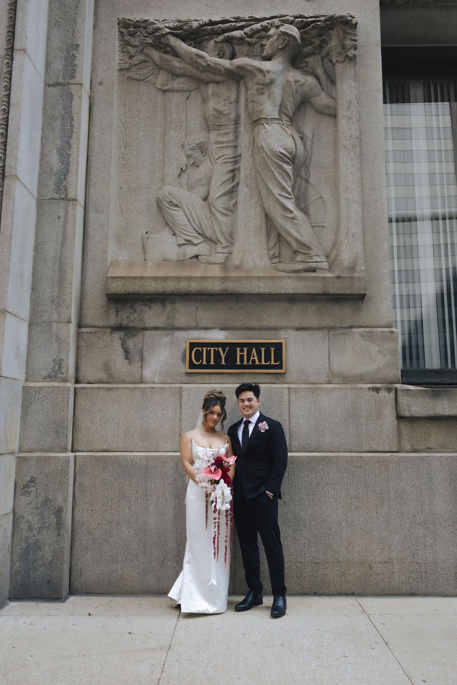 A couple in wedding attire poses against a stone wall outside City Hall for their wedding portraits.