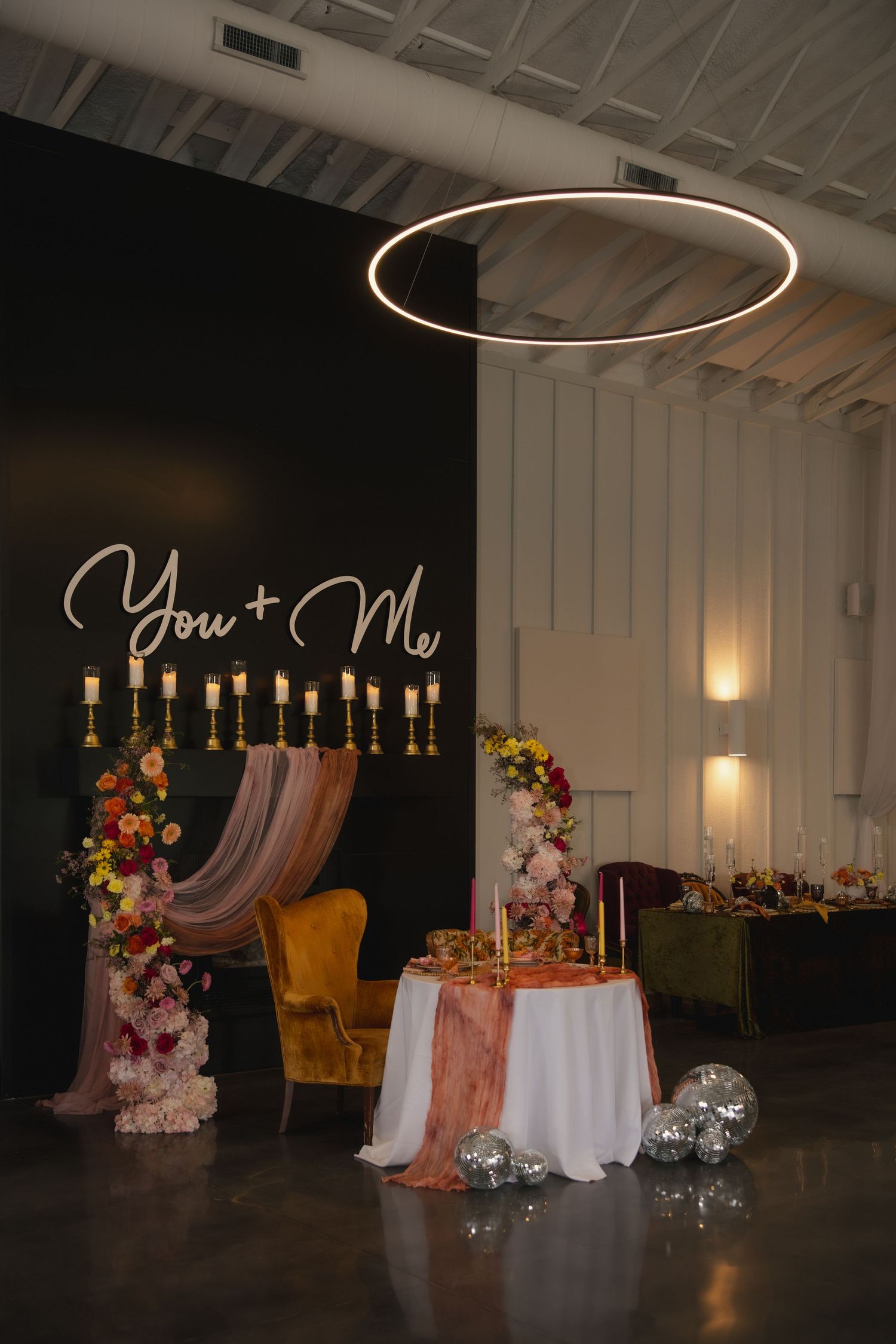 Modern wedding setup with 'You + Me' neon sign, circular ceiling light, velvet chair and floral arrangements against dark wall.