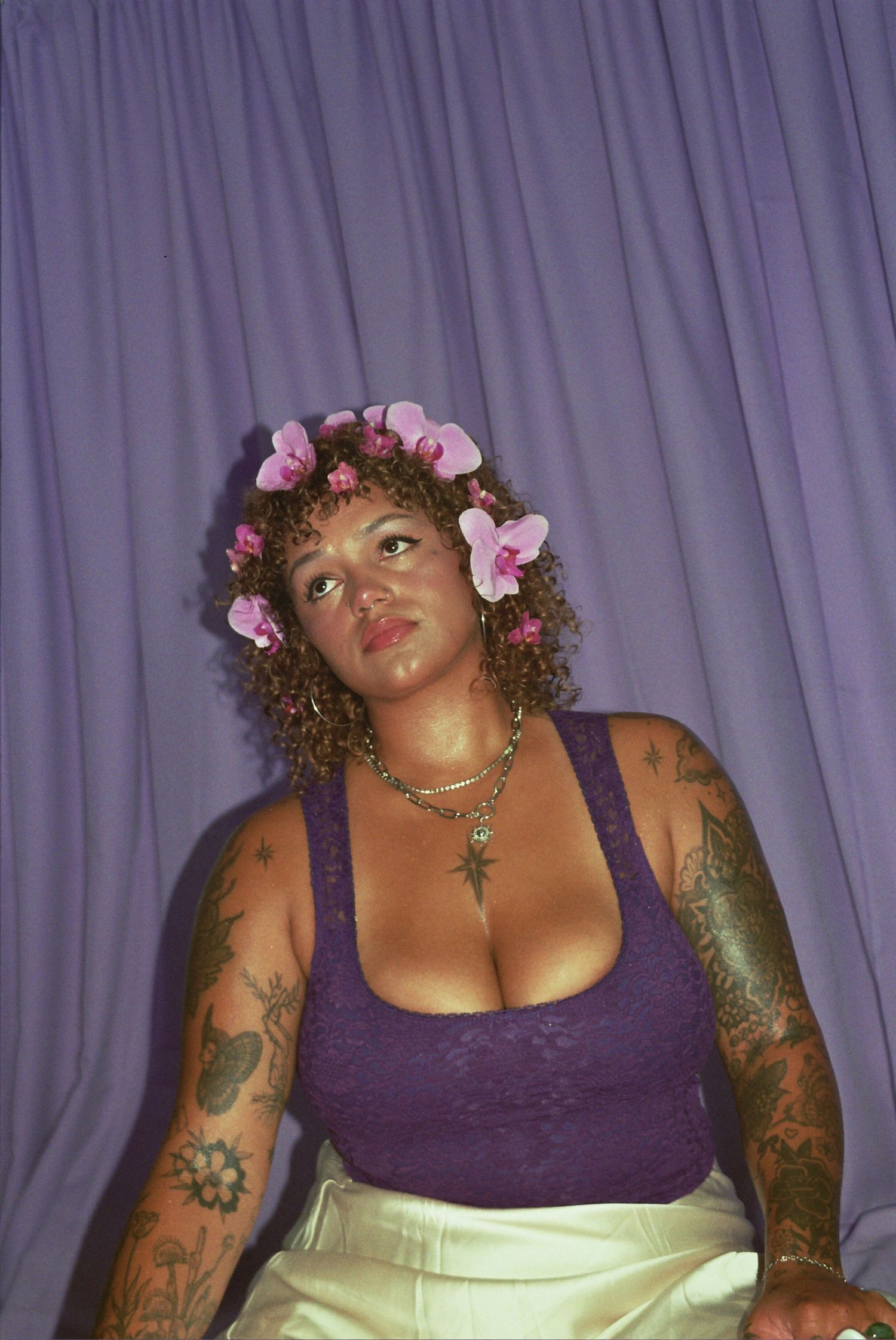 Woman with curly hair adorned with pink flowers, purple lace top, chain necklace, and tattoos against lavender backdrop.