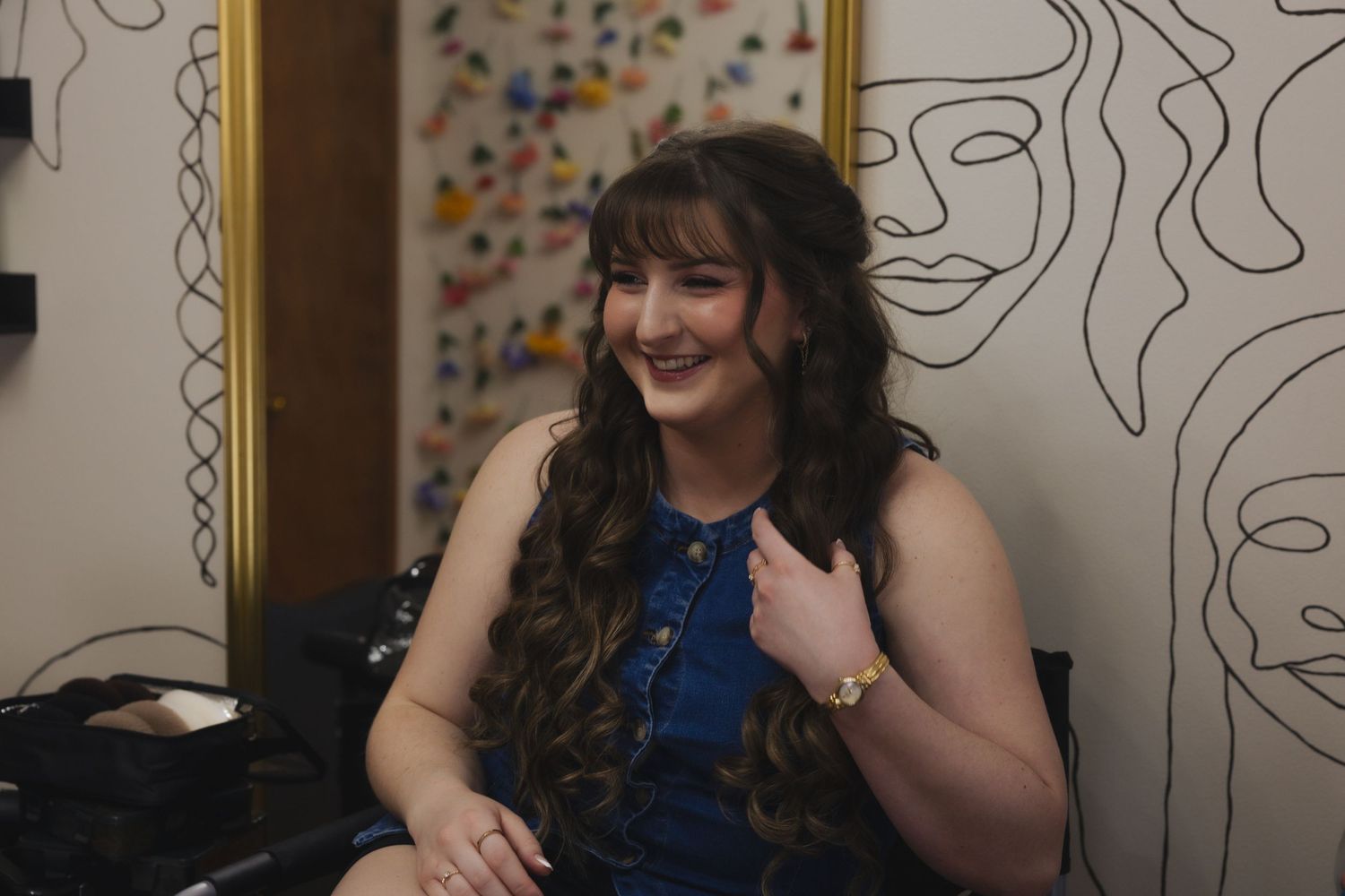 Smiling young woman with long curly hair wearing a blue dress, seated in a room with line art wall decor.