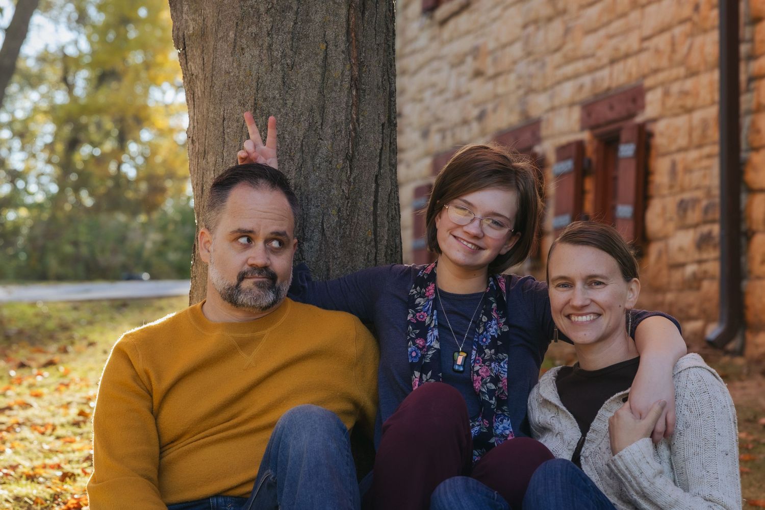 Three people sitting against a tree in autumn, smiling, with fall foliage in the background.