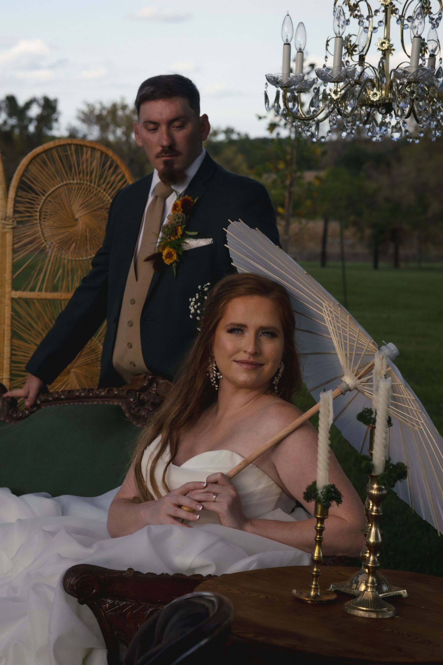 A wedding couple poses with vintage decor including a peacock chair, chandelier and candelabras in an outdoor setting.