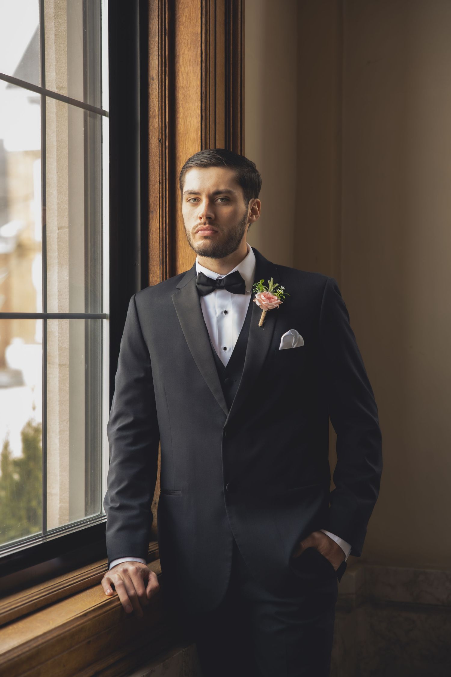 A groom in a classic black tuxedo stands by a window with a boutonniere and pocket square.