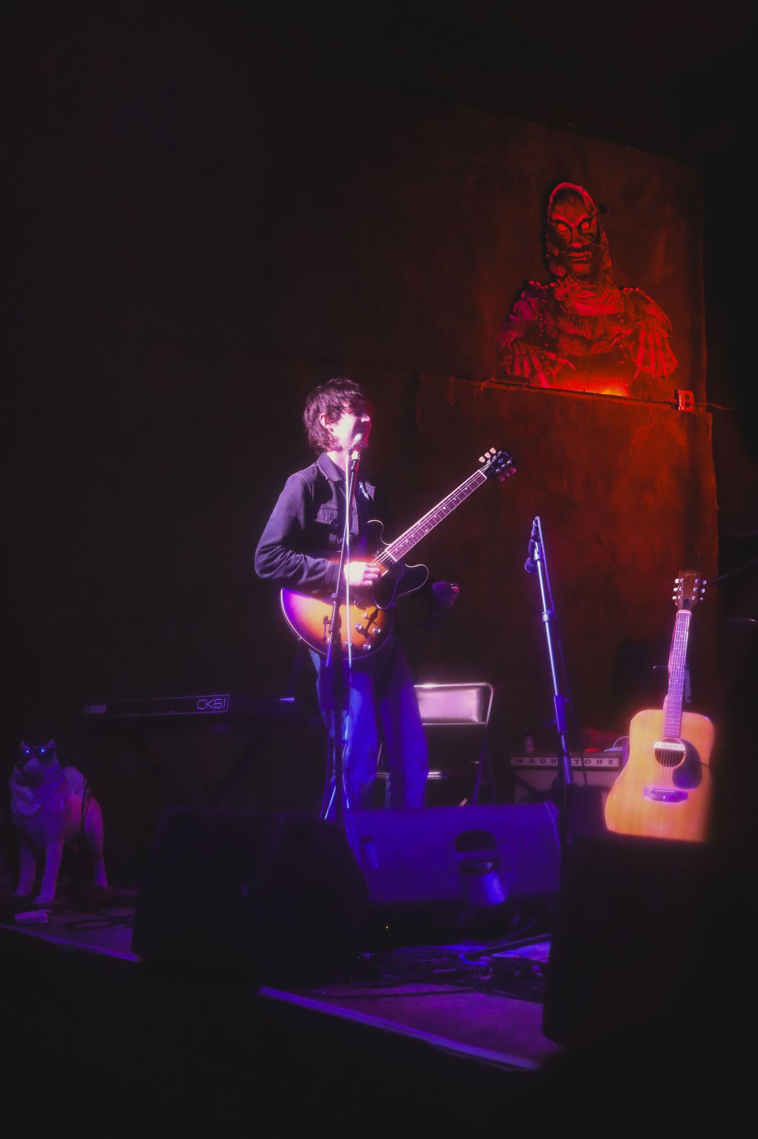 A musician performs on stage with guitars and red lighting effects casting dramatic shadows in the background.