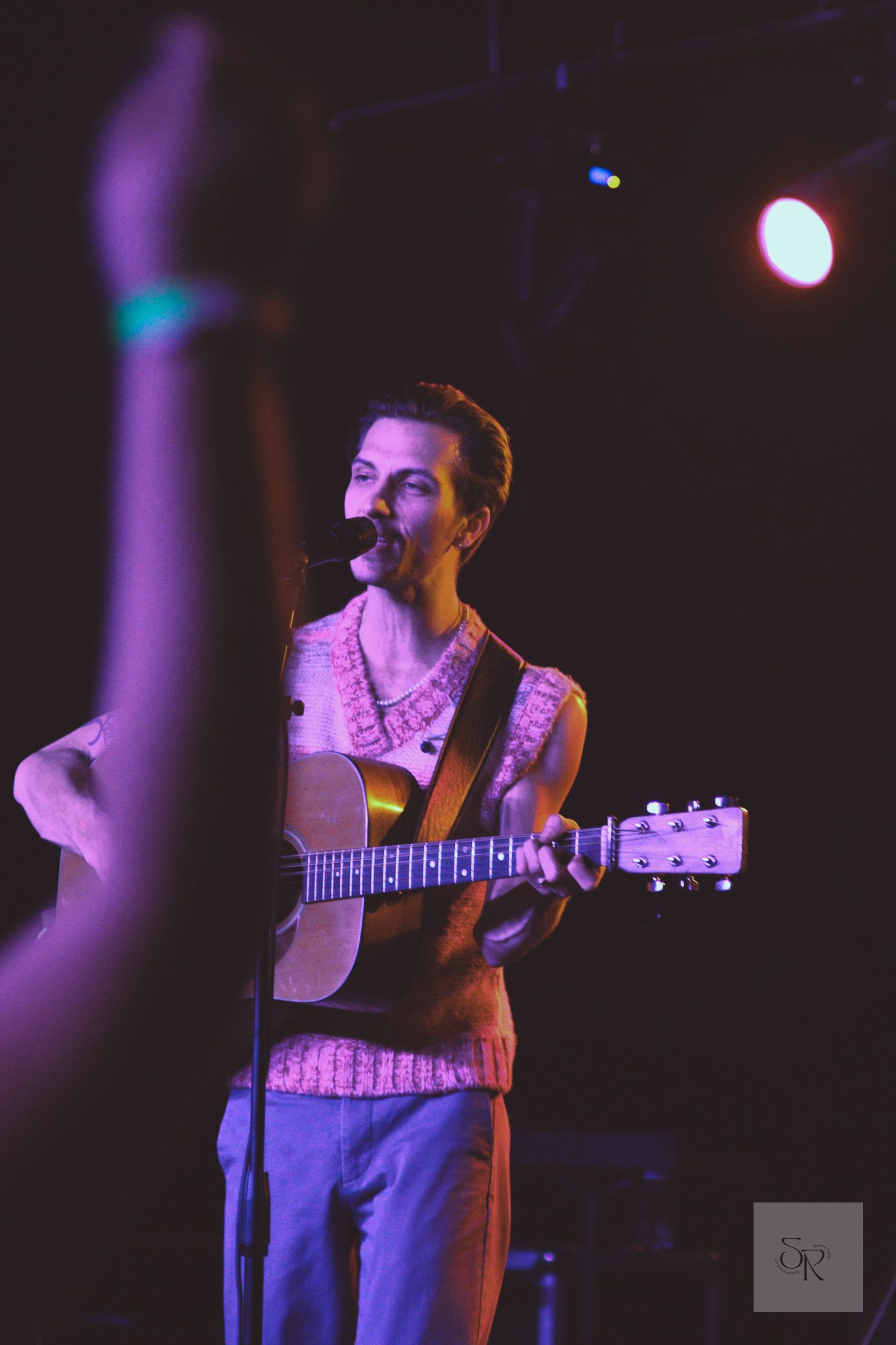 A musician performs on stage in purple lighting while playing an acoustic guitar and singing into a microphone.